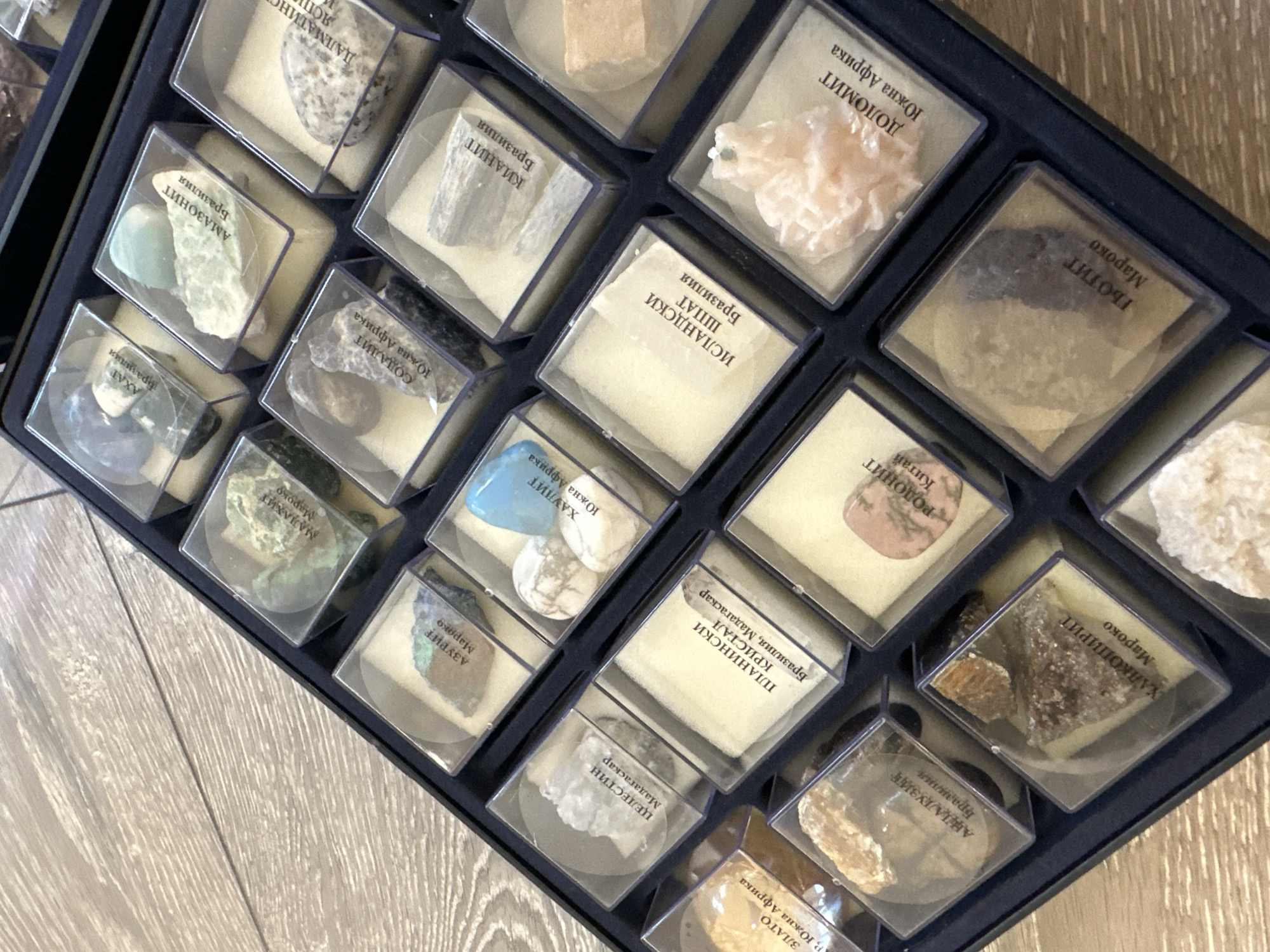 The image shows a collection of mineral samples housed in small, clear plastic cases, arranged in a dark blue, possibly plastic, tray. Each case contains a mineral specimen and a small label with text in what appears to be Cyrillic script, indicating the mineral's name and origin. The minerals vary in color and texture, including white, blue, green, pink, and brown. The tray is resting on a light-colored wooden surface.