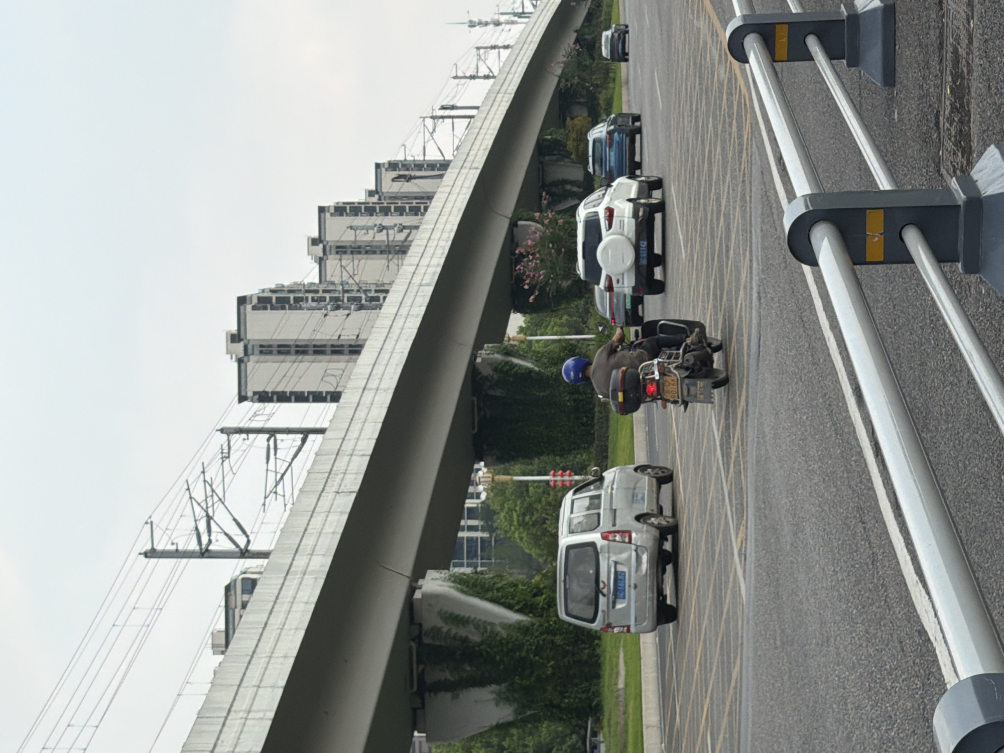 The image depicts a street scene in Jiangjin, China, under an elevated structure that appears to be a highway or railway viaduct. Several vehicles are visible on the road, including a silver minivan, a white SUV, a dark-colored sedan, a blue SUV, and a motorcycle. A person is riding the motorcycle, wearing a blue helmet and a dark jacket. The sky is overcast. The elevated structure is supported by concrete pillars, and overhead power lines are visible. Buildings resembling apartment complexes are lined up in the background. The road has lane markings and some yellow grid markings, possibly indicating a no-parking zone or a special intersection. A metal guardrail with yellow reflectors runs along the side of the road in the foreground. Vegetation, including trees and vines, grows on and around the supporting pillars of the viaduct.