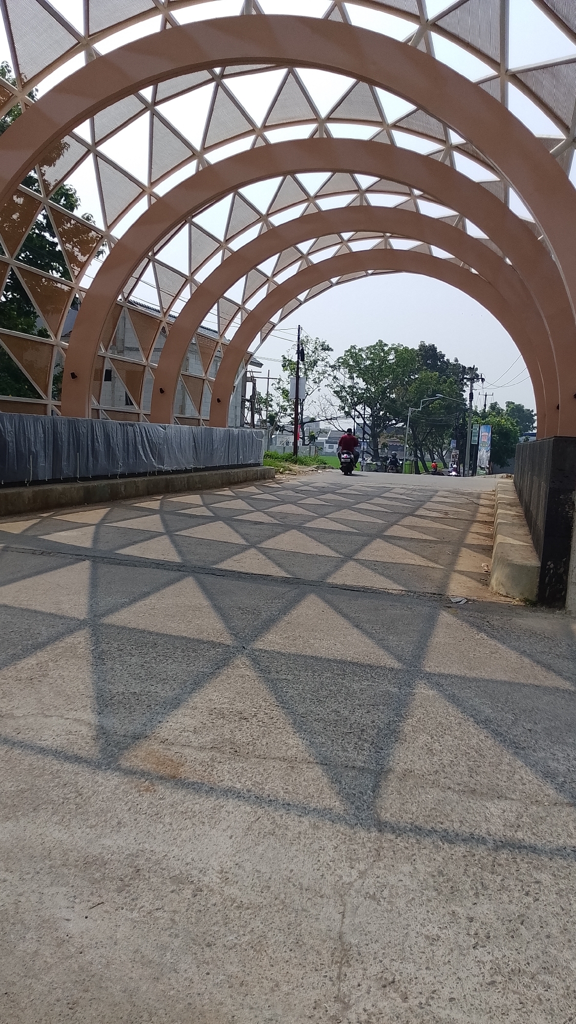 The image depicts a view through an arched walkway structure in Bojongsoang, Indonesia. The walkway's ceiling is a lattice of triangular panels, casting a geometric shadow pattern on the asphalt road below. A person wearing a red shirt is riding a dark-colored motorcycle away from the camera, entering the arched structure. In the distance, more motorcycles and trees are visible, along with buildings and utility poles. The scene appears to be a public space, possibly a pedestrian bridge or an entrance to an area.