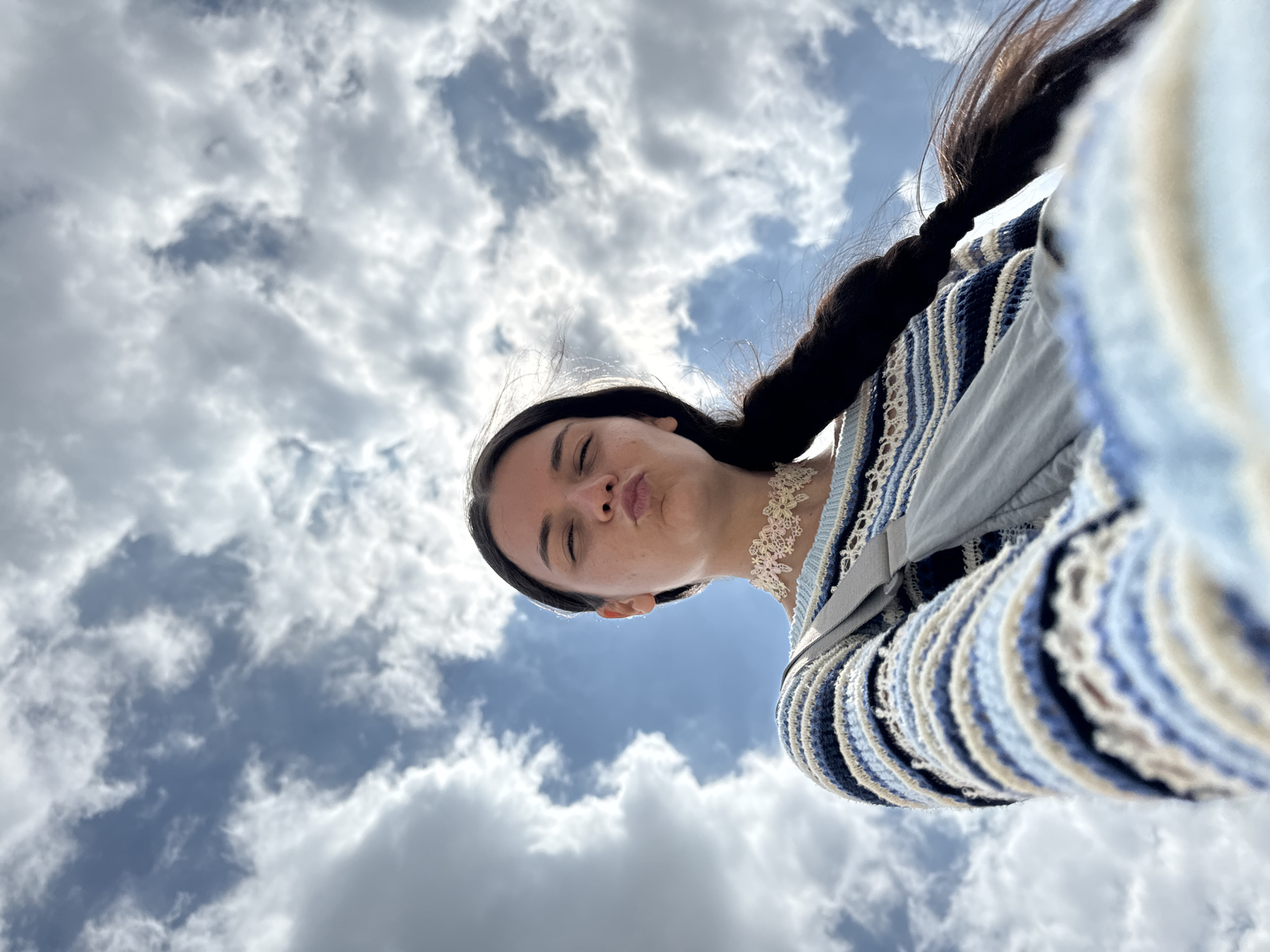 A low-angle, outdoor, medium shot captures a young woman with dark hair braided down her right side, posing for a selfie against a backdrop of a dynamic sky filled with white, fluffy clouds and patches of blue. Her face is turned towards the camera, lips pursed in a kiss. She is wearing a blue and white striped sweater with a delicate white lace choker around her neck. A pale grey bag strap is visible diagonally across her chest. The perspective suggests the photo was taken from a low vantage point, possibly lying down, looking upwards. The scene is set outdoors with a focus on the sky and the subject. There is no visible indication of the specific location within Mui Ne, Vietnam.