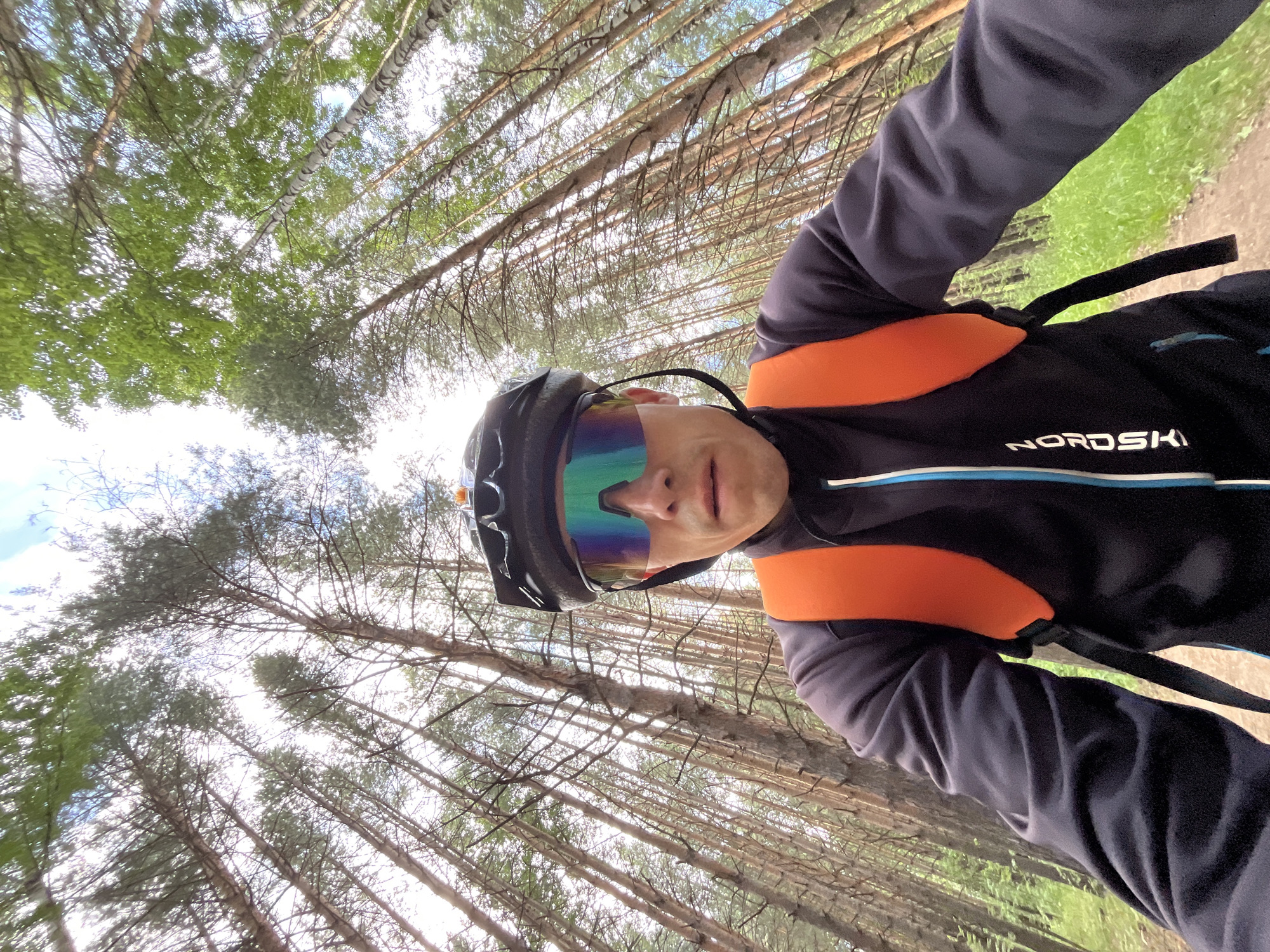 This is a first-person perspective, low-angle selfie taken outdoors, likely while cycling or hiking in a forest. The subject, a person wearing a dark blue long-sleeved shirt with orange accents and a black helmet with cycling glasses, is the primary focus. The glasses have a mirrored, iridescent lens that reflects the surrounding green foliage and sky.
The setting is a dense forest with numerous tall, slender trees, primarily pine and birch, with thin trunks and branches. The canopy of leaves is visible overhead, suggesting a temperate climate and daytime. Sunlight filters through the trees, creating a dappled effect. The ground appears to be a dirt path with patches of green grass, indicating a natural, unpaved trail.
The activity is implied to be outdoor recreation, such as cycling or mountain biking, given the protective gear worn by the subject. The upward angle of the shot emphasizes the height of the trees and the expansive sky. The image has a dynamic feel due to the low angle and the tilted perspective, common in action shots.
Visible text on the subject's shirt reads "NORDSAI" in white lettering with a thin blue line below it. The time of day appears to be daylight, possibly late morning or early afternoon, given the brightness and the angle of the light. The weather seems pleasant, with a clear sky peeking through the trees. The subject's expression is partially obscured by the glasses, but the visible portion of their face suggests a neutral or determined demeanor. The overall mood is one of an active and engaged individual enjoying nature. The location context of Chaikovsky, Russia, aligns with the type of forest depicted.
