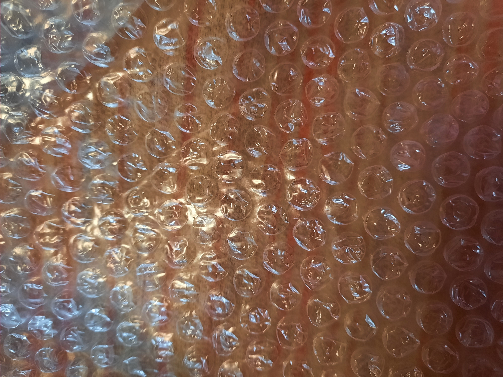 The image displays a close-up, overhead view of bubble wrap. The transparent plastic bubbles are arranged in a honeycomb pattern, with some appearing more inflated than others. The background, visible through the translucent bubbles, is a warm-toned, possibly wooden surface with some reddish-brown striations, suggesting a table or a floor. There are no people, actions, or interactions visible. The lighting creates highlights and shadows within the bubbles, adding depth and texture to the surface. The image does not contain any information that would allow for the determination of its location in the city of Rosenheim, Germany.