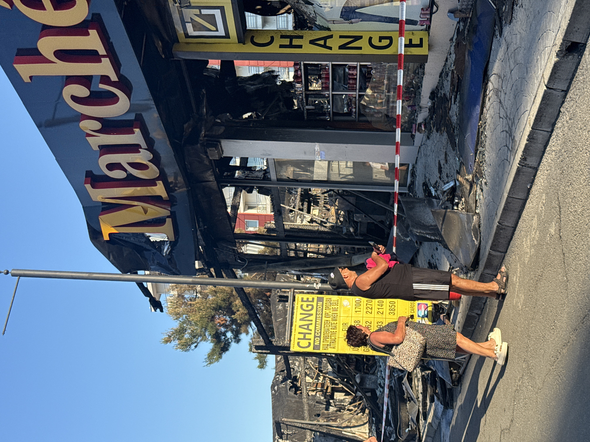 The image captures a street scene in Nesebar, Bulgaria, with a focus on a building that appears to have been damaged by fire. The storefront of a business named "Marche" is prominently displayed, with its large gold and red lettering intact despite the surrounding destruction. The facade of the building is charred and blackened, with debris scattered on the pavement in front.
To the left of the Marche store, a yellow sign with black text reads "CHANGE NO COMMISSION" and displays currency exchange rates for EUR, GBP, and USD. Two individuals, a woman and a man, are standing near this sign, observing the scene. The woman, wearing a patterned dress and carrying a leopard-print bag, stands with her back to the camera. The man, in a black tank top and shorts, is facing towards the woman and looking at his phone.
A red and white striped tape is stretched across the area in front of the building, indicating a restricted zone. Beyond the immediate storefront, the interior of the Marche store is visible, showing shelves with various items, some of which are obscured by smoke and fire damage. The overall environment suggests a post-fire scene on a sunny day, with a clear blue sky overhead and trees visible in the background.