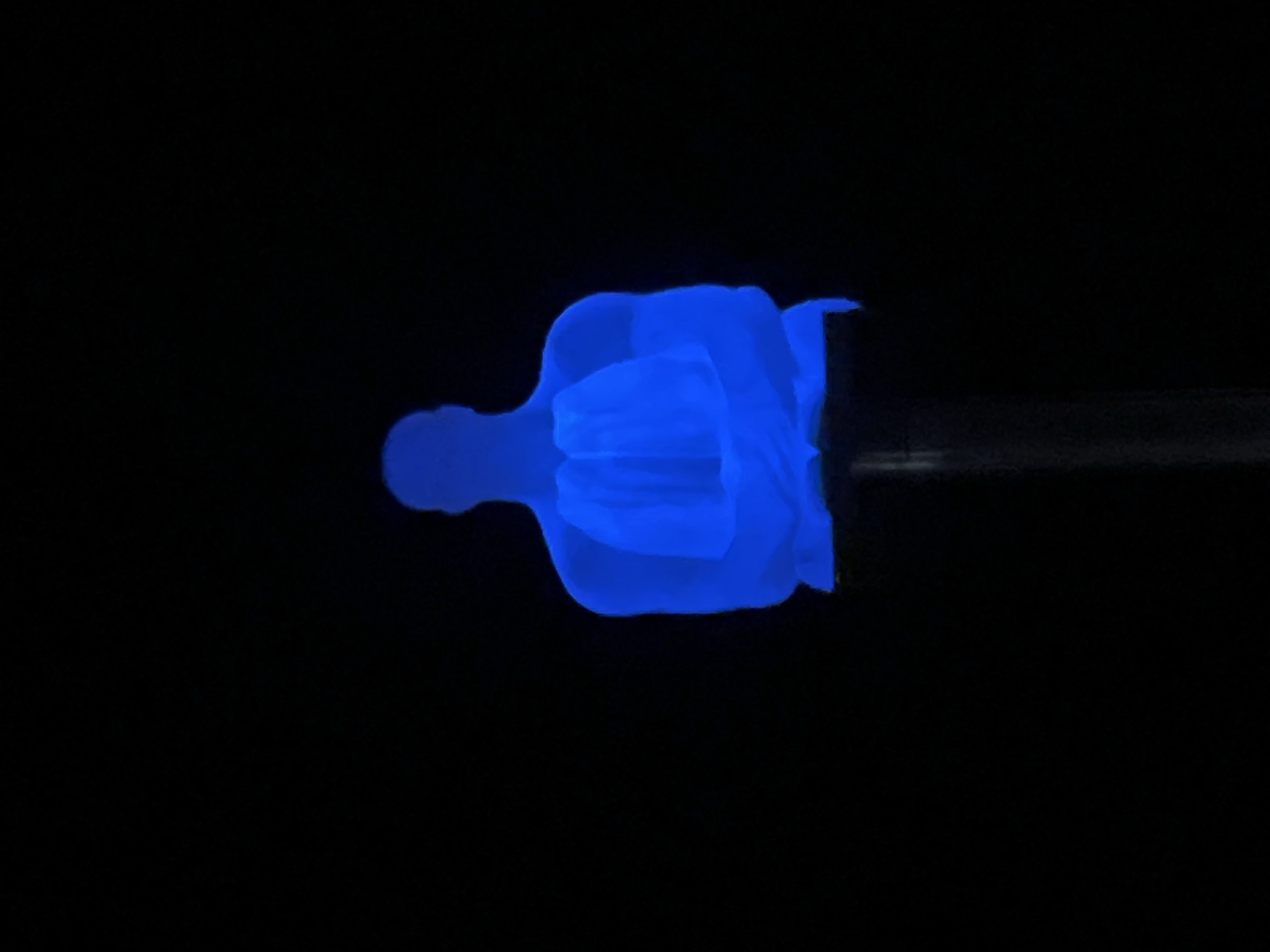 A figure, possibly a person, is illuminated in blue against a completely black background. The figure is oriented horizontally, with what appears to be the head to the left. The body and limbs are distorted or abstracted, giving the impression of movement or transformation. A cylindrical, semi-transparent object extends from the right side of the figure. The environment is entirely dark, providing no context for location. It is not possible to identify the city of Yerevan, Armenia, or any specific setting.