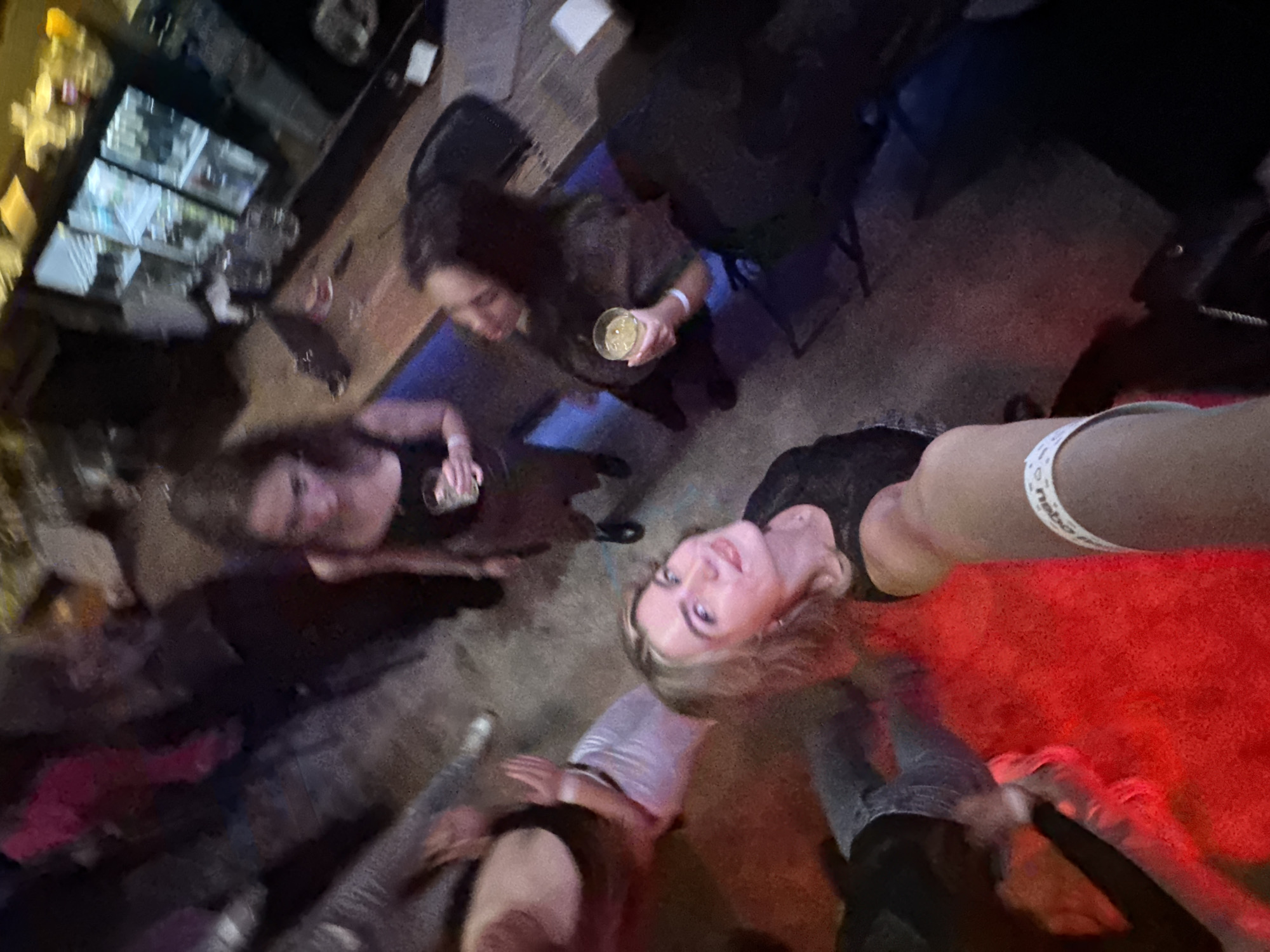 Four women are gathered in a room with dim lighting, likely a bar. One woman is holding a glass of liquid. The room appears to have a bar counter and a table with chairs. The city is Unknown, Unknown.
