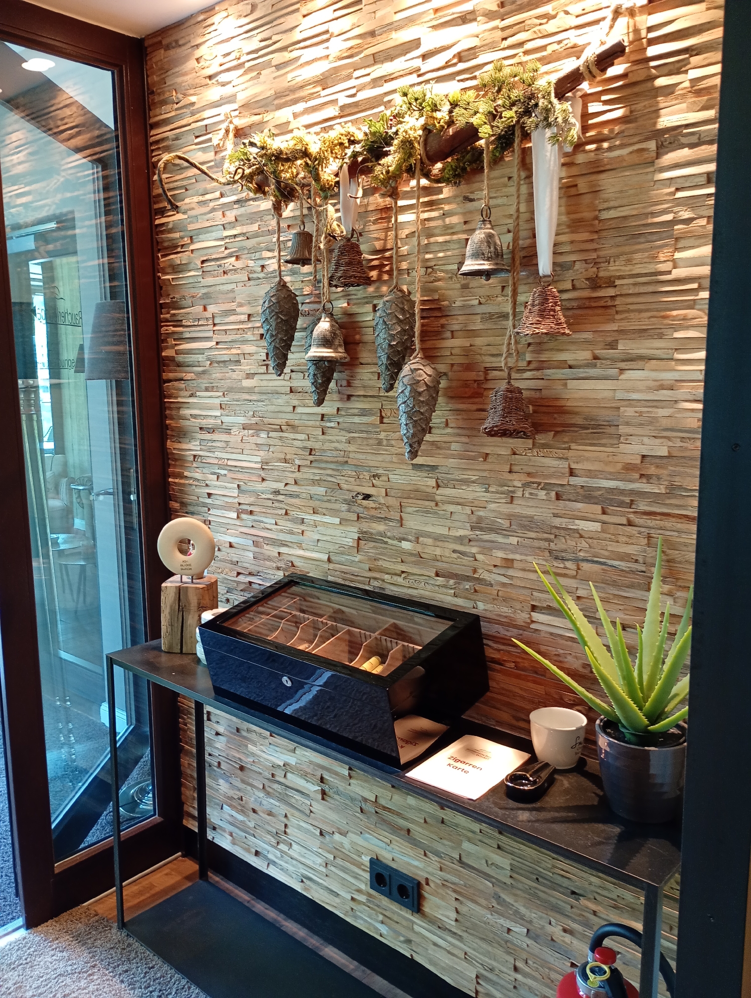 This image depicts an interior scene, likely a lounge or reception area, characterized by a textured wooden panel wall. The wall is adorned with decorative elements, including hanging pinecones, bells, and greenery suspended from a branch. Below the wall adornments, a dark console table holds several items. Prominently displayed is a dark, rectangular box with a glass lid, revealing dividers within, possibly for storing cigars or other small items. Adjacent to this, a small white cup, an ashtray, and a folded card are visible. To the right, a potted aloe vera plant stands beside the console. A tall, dark vertical element frames the right side of the image, and a glass door or window is situated on the left, offering a partial view of an interior space with furniture. The overall impression is one of a well-appointed, perhaps rustic-themed, indoor setting. The text on the card on the console reads "Zigarren Karte", which translates to "Cigar Menu", and the location is identified as Bad Sachsa, Germany.
