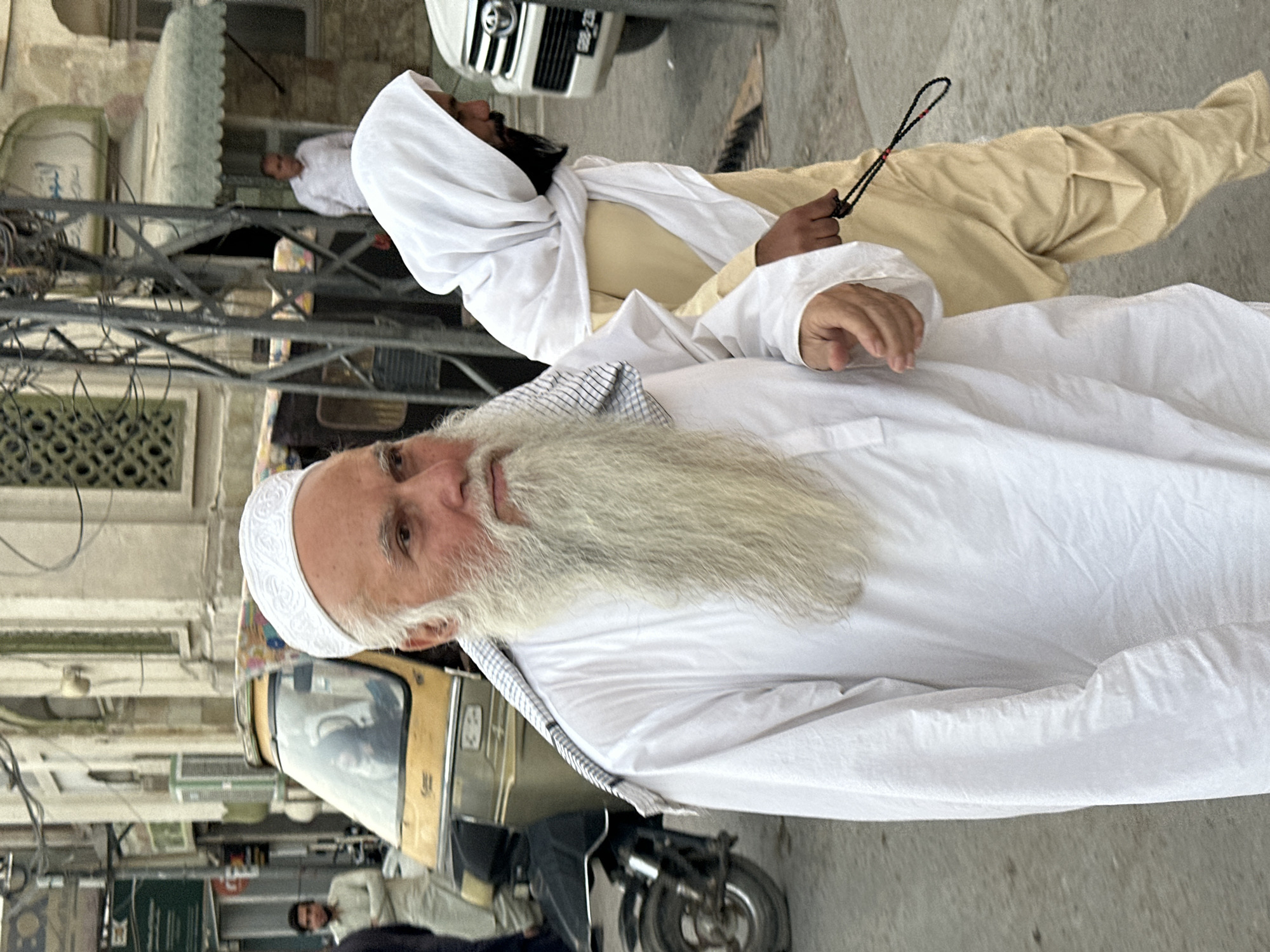 In Quetta City Tehsil, Pakistan, two adult males are walking on a paved street. The man in the foreground, on the left, is wearing a white embroidered cap and a long-sleeved white tunic, with a black and white checkered cloth draped over his left shoulder. He has a long, grey beard and is looking towards the left of the frame. The man on the right is wearing a light-colored headscarf and a light-colored tunic and trousers. He is holding a string of black beads in his right hand and looking towards the right of the frame. In the background, to the left, a yellow auto-rickshaw is visible, with a person seated inside. Further in the background, a white SUV is parked on the right. The environment appears to be an urban setting with buildings lining the street.