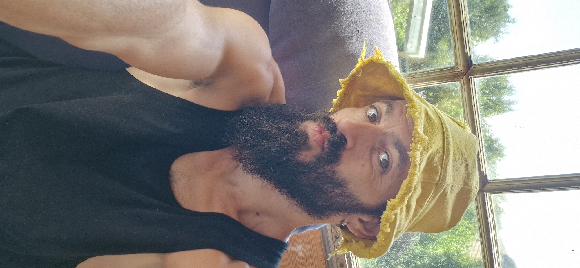 A man with a long, dark beard and a yellow bucket hat is shown from the chest up, looking towards the camera. He is wearing a black tank top and has a dark grey chair behind him. Sunlight streams through a large window behind him, with trees and greenery visible outside. The image is a selfie, taken indoors, likely in a residential setting in Wattenwil, Switzerland.