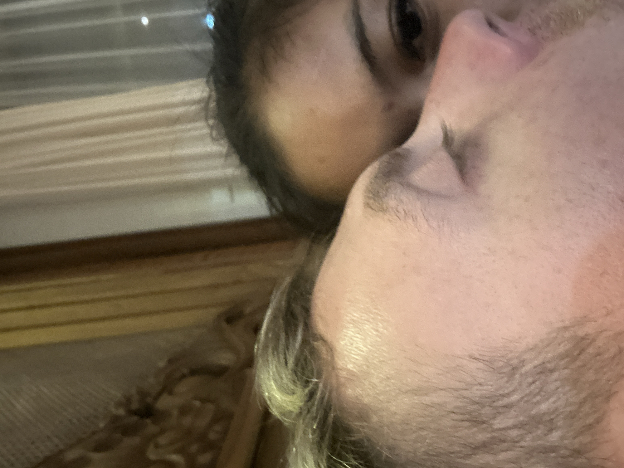 A close-up shot of two individuals, one appearing to kiss the other. The person on the left has dark hair and one visible eye, while the other individual has light hair and closed eyes. The setting includes window blinds and appears to be indoors. There is no specific information available to confirm the location in Ubud District, Indonesia.