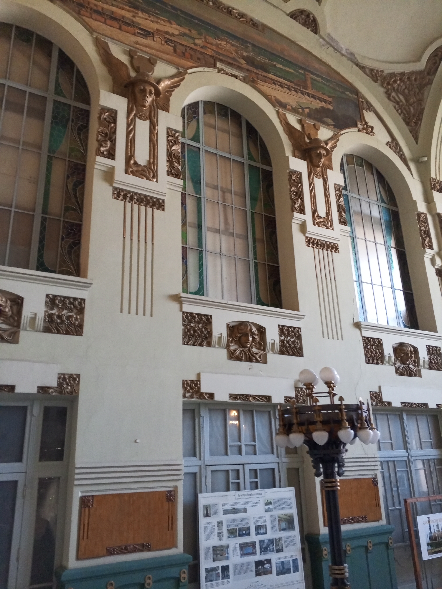 The image depicts the interior of the main hall at the Vitebsky railway station in Saint Petersburg, Russia. The scene showcases a high, pale cream-colored wall extensively adorned with Art Nouveau architectural and decorative elements.
Three large arched windows, featuring gridded panes and subtle stained-glass accents in greens and blues, dominate the upper wall. Above these windows, a lengthy mural depicts an industrial or urban landscape in muted tones.
Between the two rightmost arched windows, a prominent gold-toned relief sculpture of a winged male bust wearing a helmet (resembling Mercury/Hermes) is centered within a vertical decorative panel, flanked by smaller floral reliefs. Below the windows, at a lower level, multiple square gold-toned relief panels feature female faces with flowing hair and additional floral patterns. Vertical gold-toned strips and intricate decorative motifs are present in the arch spandrels and along the ceiling lines.
In the lower right foreground, a large, ornate lamppost with a dark base and multiple spherical white lamps with gold accents is visible. Adjacent to it, a white information display board containing text and numerous small photographs, likely detailing the station's history or features, stands alongside a simpler wooden-framed display. No individuals are present in the scene.