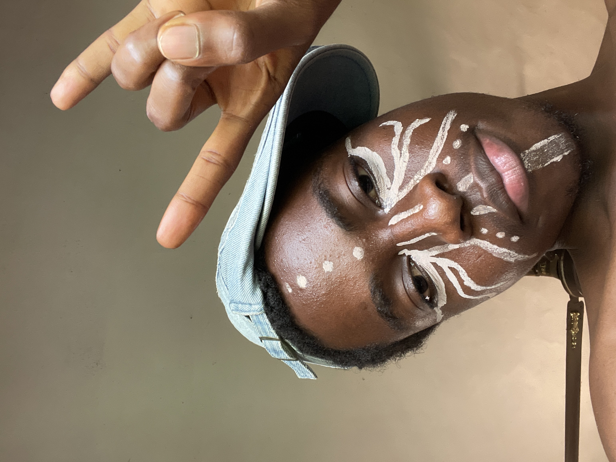 An African male wearing a denim cap with white painted markings on his face is angled towards the left with his body slightly sideways. The man is making a hand gesture with the pointer and pinky fingers extended, with his ring and middle fingers folded towards the palm. The environment seems to be indoors with a plain neutral background. A strap attached to what appears to be a handbag or shoulder bag is visible. There is no visible location context to suggest Nkpor, Nigeria.