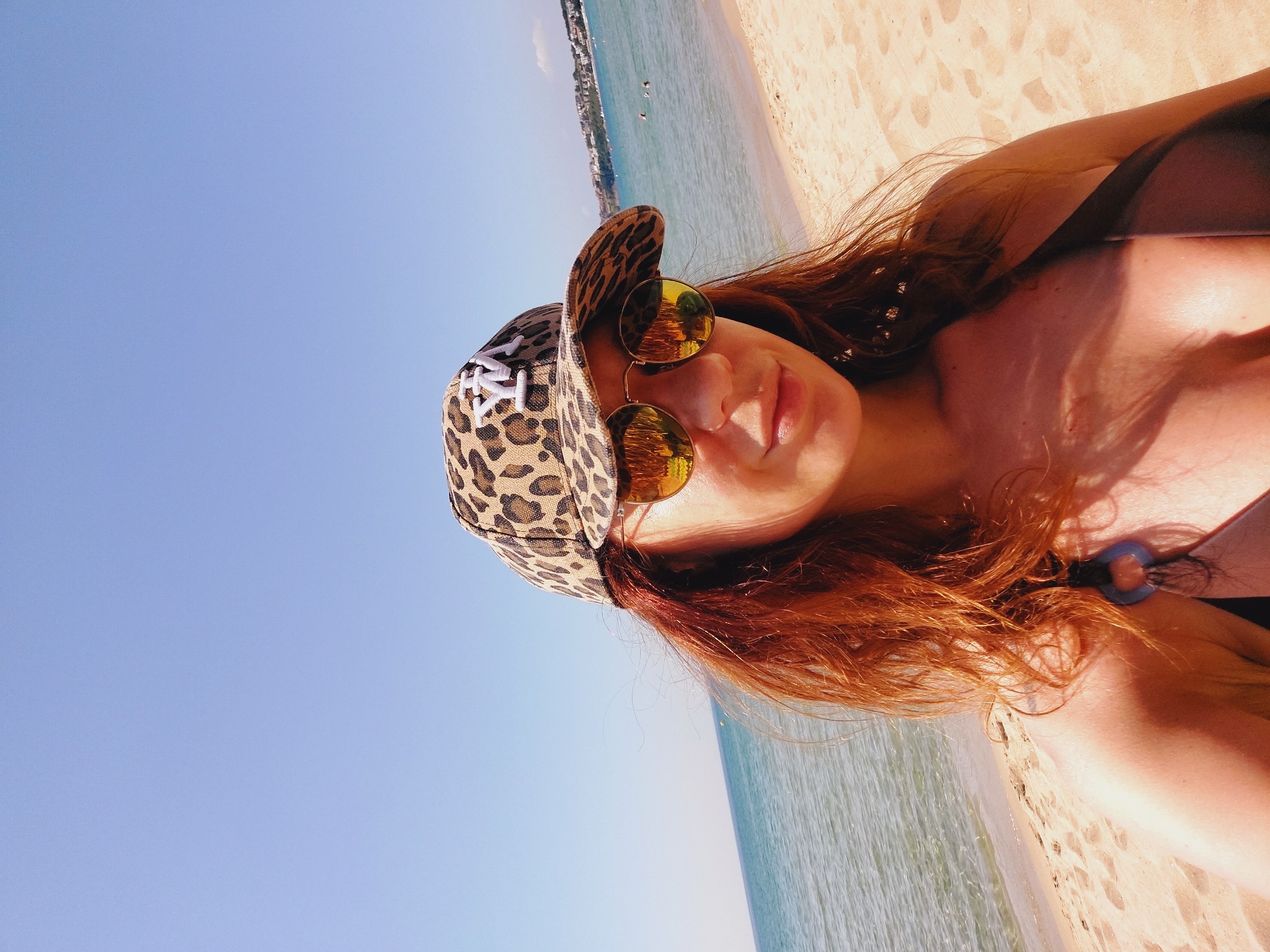 A woman with long, reddish-brown hair is standing on a sandy beach, with the sea and a clear blue sky behind her. She is wearing a leopard-print baseball cap with a white logo, and reflective sunglasses. She is also wearing a dark-colored bikini top. The woman is looking towards the camera and smiling slightly. The beach appears to be located in Sozopol, Bulgaria, as indicated by the distant view of buildings and coastline typical of the area. The water is calm, and there are a few other people visible in the distance, swimming or wading in the sea. The sun is shining brightly, casting shadows on the sand and the woman's face.