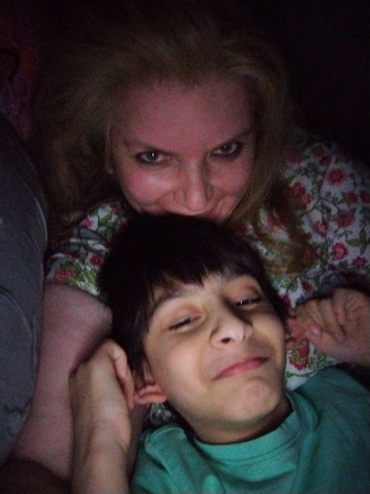 A dimly lit, close-up photograph captures two individuals in a vertical composition. An adult female with blonde hair is positioned at the top, looking directly into the camera with open eyes and a subtle smile. She wears a floral patterned garment featuring red, green, and white elements. Below her, a male child or young adolescent with dark hair and bangs is partially reclined, his eyes partially closed in a smile. His right hand is raised near his ear. He wears a solid green long-sleeved top. A dark, possibly grey, fabric item is visible on the far left. The scene is characterized by low light, casting soft illumination on the subjects' faces against a dark, indistinct background, suggesting an indoor setting. This image is associated with the city of Al Mas'oudi, United Arab Emirates.