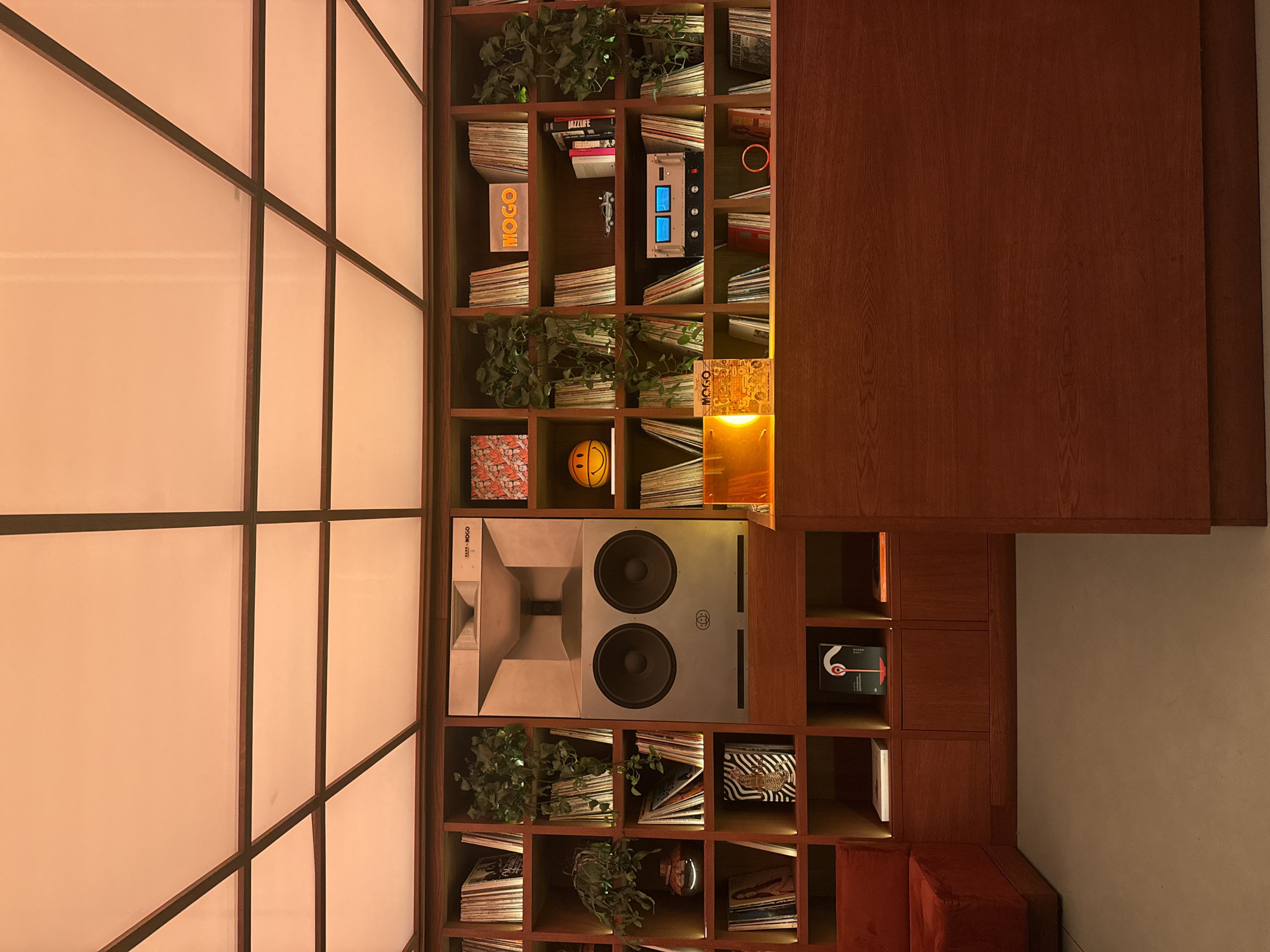 An interior shot captures a stylish and well-organized space, likely a lounge or common area, with a large, custom-built wooden shelving unit serving as a central feature. The shelving unit occupies the majority of the background, filled with numerous compartments.
Many shelves are stacked with books or vinyl records, oriented horizontally, suggesting a curated collection. Green potted plants are interspersed throughout the shelves, adding a touch of nature. Prominently displayed is a large, contemporary speaker system in a gray finish, featuring two large circular woofers and a distinctive horn-shaped element, with the text "MARK MOGO" visible on its side. Above it, another piece of audio equipment, silver-toned with two glowing blue screens and various controls, is integrated into the shelving.
Other decorative elements include a bright yellow block spelling "MOGO," a glowing yellow sign also reading "MOGO," and a yellow smiley-face ball. A book titled "JAZZ LIFE" is visible, along with other items like a small silver toy car and an orange ring. The shelves are illuminated by warm, integrated lighting, creating an inviting ambiance.
To the left of the shelving, a translucent partition with a dark grid pattern resembles a modern shoji screen, allowing soft, diffused light to pass through. To the right, a large, plain wooden panel matches the tone of the shelving. A segment of a red, upholstered seating area is visible in the bottom right corner. The warm, artificial lighting suggests an evening setting in this modern, design-focused space located in Milan, Italy.