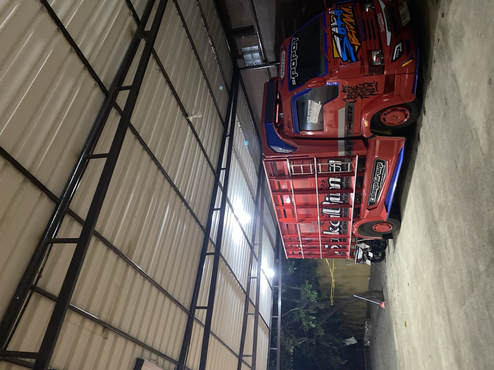 The image shows a bright red truck parked indoors under a corrugated metal roof supported by a dark metal frame. The truck has blue accents on its cab and a large cargo box decorated with red lettering and graphic designs, including the words "Sikocillpim" and "Cahaya Group." A motorcycle is parked behind the truck's cab. To the left of the truck, a broom leans against a tree. The ground is paved and appears to be a concrete or asphalt surface. The scene is illuminated by overhead lights. The overall impression is of a vehicle in a storage or workshop area, potentially in a more industrial or commercial part of Tangerang, Indonesia, given the vehicle's appearance and the common use of such structures for vehicle storage and repair in the region.
