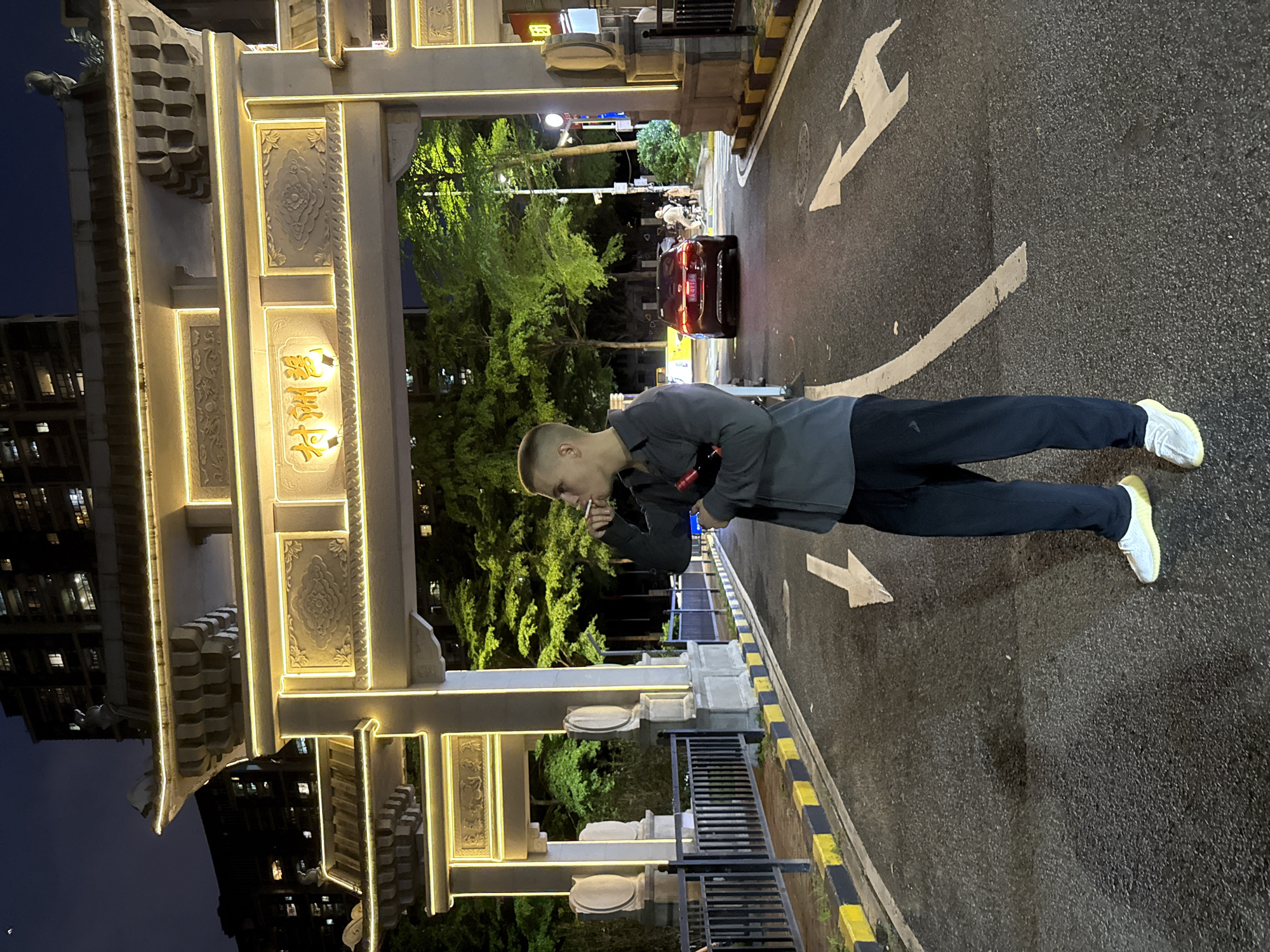 The image depicts a nighttime urban scene in Haizhu District, China, featuring a prominent, brightly illuminated traditional Chinese archway.
In the foreground, slightly right of center, a young adult male stands on a wet asphalt road. He has a very short buzz cut, wears a dark grey long-sleeved shirt, and dark blue trousers with white sneakers that have light yellow soles. He holds a dark red bottle tucked under his left arm and raises his right hand to his mouth, holding a lit cigarette or vape from which smoke or vapor is exhaling. He gazes towards his left.
Behind the man is a large, multi-tiered archway or gate, typical of traditional Chinese architecture, featuring decorative carvings and roof sculptures. The archway is outlined with bright yellow LED lights, illuminating Chinese characters on its central panel, which read "善洲村" (Shanzhou Village). Two smaller archways flank the main one.
The road features white painted arrows indicating traffic direction and a solid white center line. Both sides of the road have black and yellow painted curbs and dark metal railings. Beyond the railings, green trees are visible, some brightly illuminated from below.
In the background, a dark-colored sedan with illuminated taillights (license plate 粤A.4T168) is either parked or moving slowly away from the viewer. Further back, on a sidewalk, several indistinct human figures are visible. Tall, dark buildings with scattered illuminated windows rise in the distant background under a dark night sky, with a sliver of the moon visible in the upper left corner.