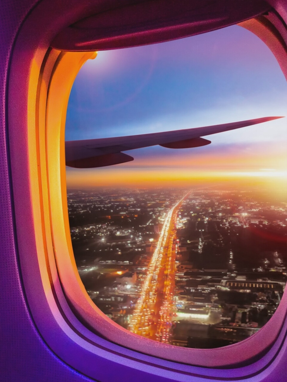 The image shows a view from an airplane window overlooking a city at sunset. The airplane's wing is visible in the upper right portion of the frame. Below, a long, illuminated highway cuts through the city, with streaks of car lights suggesting heavy traffic. The city itself is a tapestry of twinkling lights against a darkening landscape. The window frame is illuminated with warm, gradient lighting, transitioning from purple to orange. The sky above is a blend of blue and orange, characteristic of a sunset. The scene is of an urban environment during twilight, viewed from an aerial perspective. There is no indication of French Pass / Anaru, New Zealand.