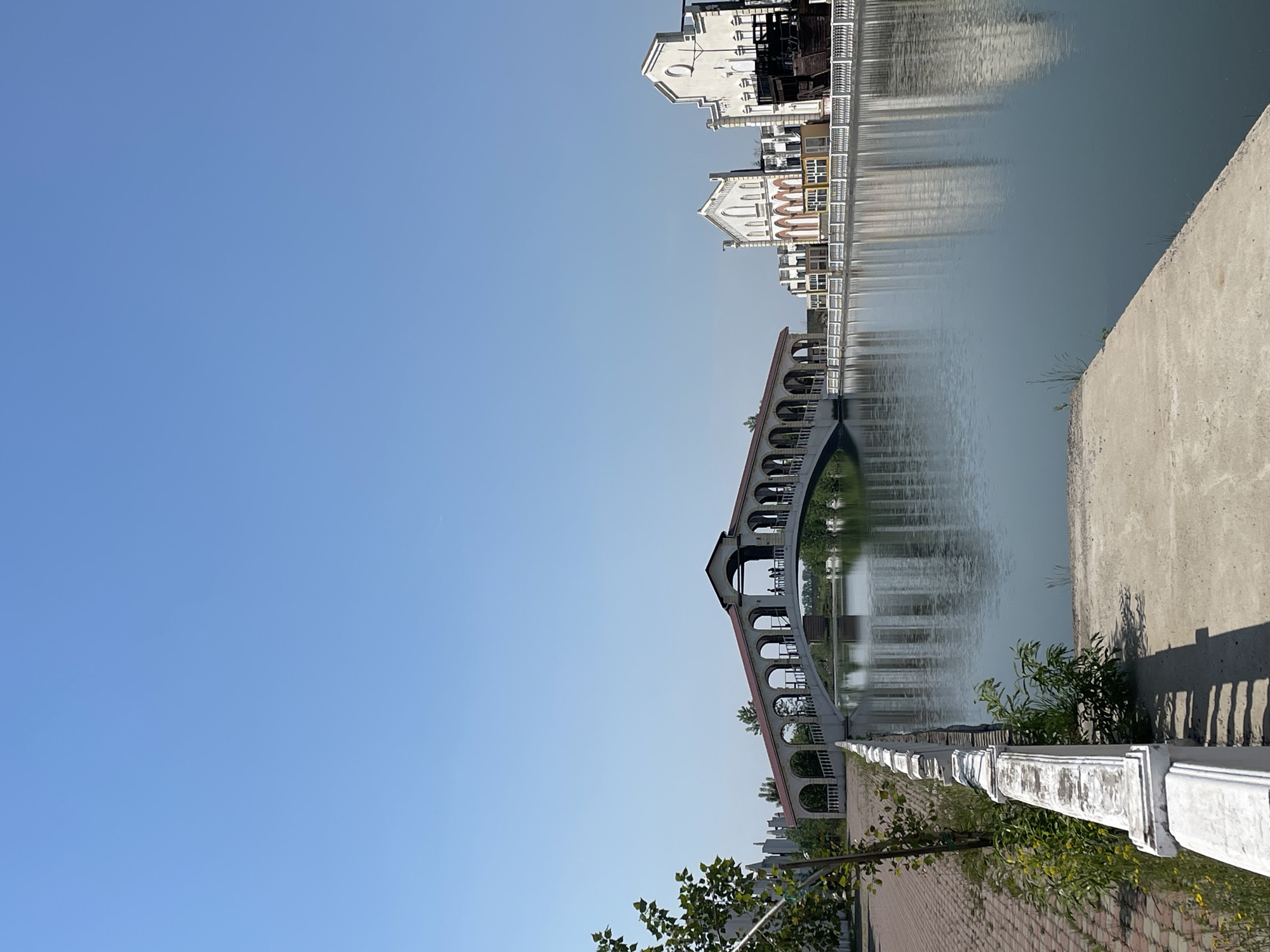 This image depicts a wide outdoor scene in the city of Unknown, Russia, under a clear, bright blue sky. A calm body of water, likely a canal or river, flows across the middle ground, reflecting the sky, structures, and vegetation with visible distortion.
Spanning the water in the left-center is a prominent, arched bridge, likely constructed from light-colored stone or concrete, featuring a reddish-brown roof over its central section. The bridge incorporates multiple arched openings and a white balustrade along its pedestrian path. Several dark, human silhouettes, estimated to be three to four individuals, are visible walking across the bridge.
On the far bank, to the right of the bridge, a series of light-colored buildings with distinct architectural features are visible. These structures, primarily white or cream with some reddish-brown accents, feature elements such as arches, gables, and decorative facades, suggesting a themed architectural style. One building on the far right has an integrated or adjacent dark, wooden, ship-like structure.
The near bank in the foreground features a reddish-brown brick paved walkway on the left, bordered by a white balustrade running parallel to the water's edge. This balustrade shows signs of weathering and discoloration. Green foliage, including a small tree with prominent leaves, grows along the walkway and near the balustrade. On the right side of the foreground, a concrete or paved area extends towards the water, sloping gently downwards. Shadows cast across the concrete and brick paths indicate a sunny day.