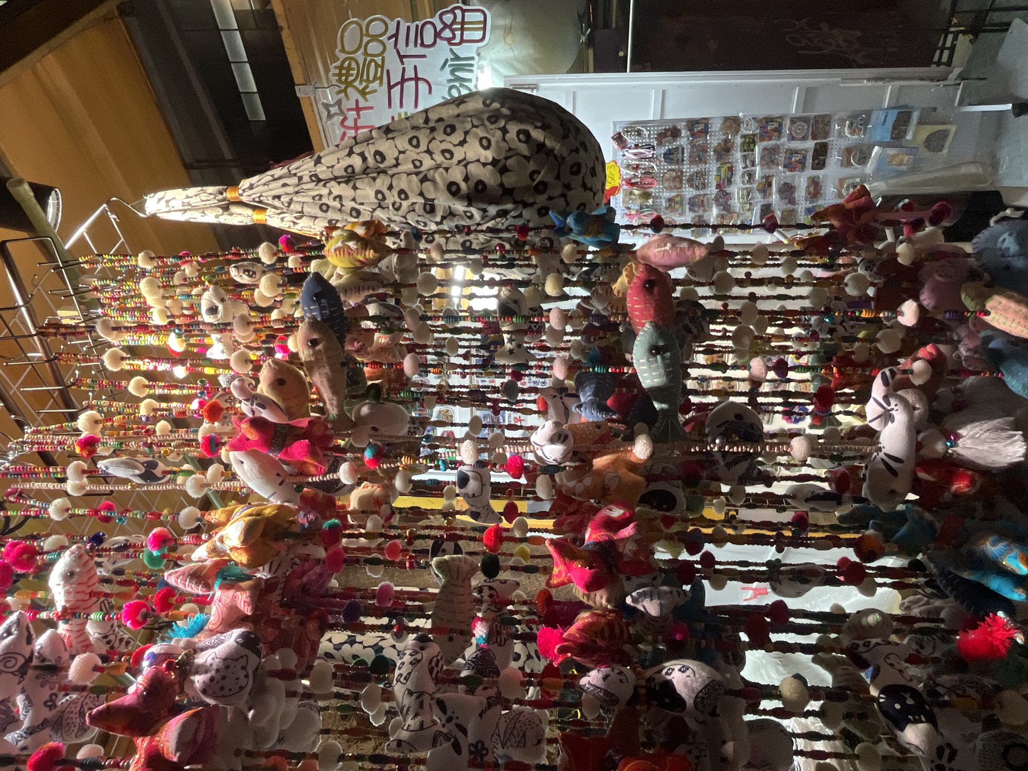 The image shows a street market stall in Hong Kong, China, displaying numerous handmade fish-shaped ornaments hanging from strings with beads. The ornaments are made of various fabrics, with different patterns and colors. Above the stall, there are printed signboards, and further back, a wall with additional merchandise. The stall is densely packed, illuminated by artificial lighting.