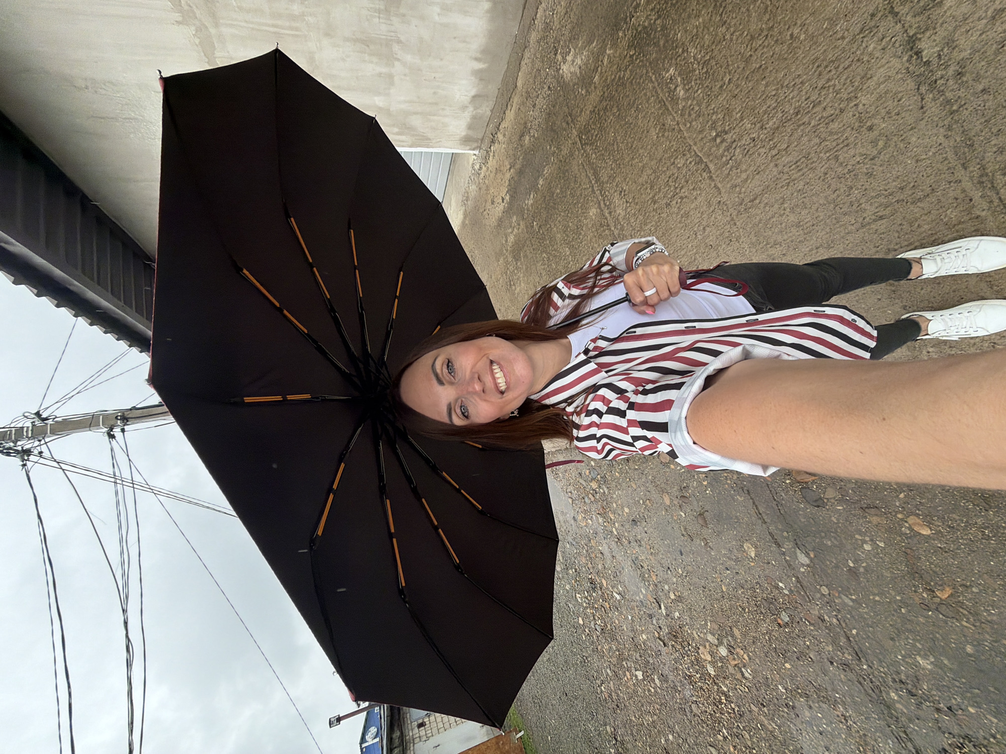 A woman is taking a selfie while holding a large black umbrella over her head. She is wearing a striped shirt, white t-shirt, black pants, and white sneakers. The background shows a dirt and gravel path, a building with a concrete wall, and a utility pole with wires. The sky is overcast. The location is in Birobidzhan, Russia.