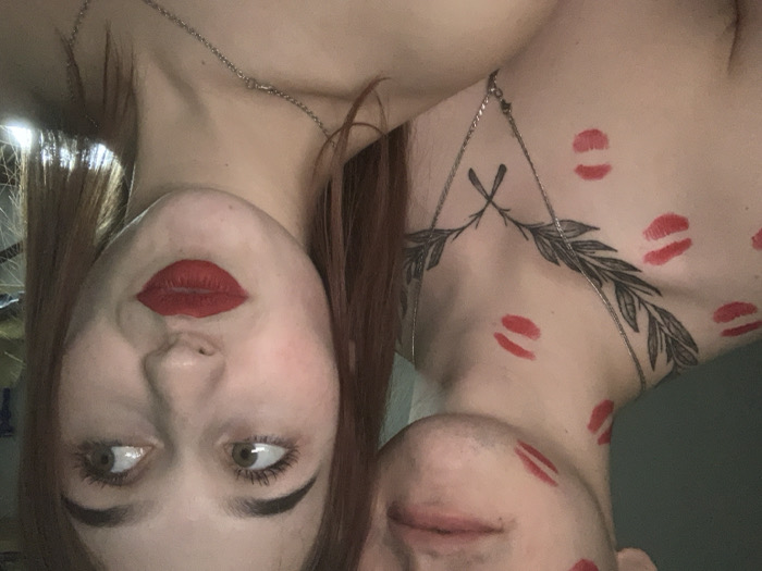 The image displays a close-up shot of two individuals, presented upside down due to a 90-degree counter-clockwise rotation.
On the left side of the frame (when viewed correctly, this would be the top), a person with reddish-brown hair, likely a woman, is visible. She has dark, well-defined eyebrows and bright red lipstick on her lips, which are slightly pursed. Her eyes are open and looking upwards and slightly to her left. A thin silver chain necklace is visible around her neck. Several red lipstick marks are visible on her neck and shoulder area.
On the right side of the frame (when viewed correctly, this would be the bottom), the upper chest and lower face of another person are shown. This individual appears bare-chested and has a prominent black tattoo on their upper chest, which depicts a symmetrical design resembling a laurel wreath or intertwined leafy branches. A similar thin silver chain necklace is draped over their chest, crossing over the tattoo. Their chin and lower lip/mouth area are visible, also adorned with multiple red lipstick marks, similar to those on the first individual.
The overall scene suggests an intimate and playful moment between the two, characterized by the scattered lipstick kisses on their skin. The lighting appears to be indoor. No text is visible in the image.