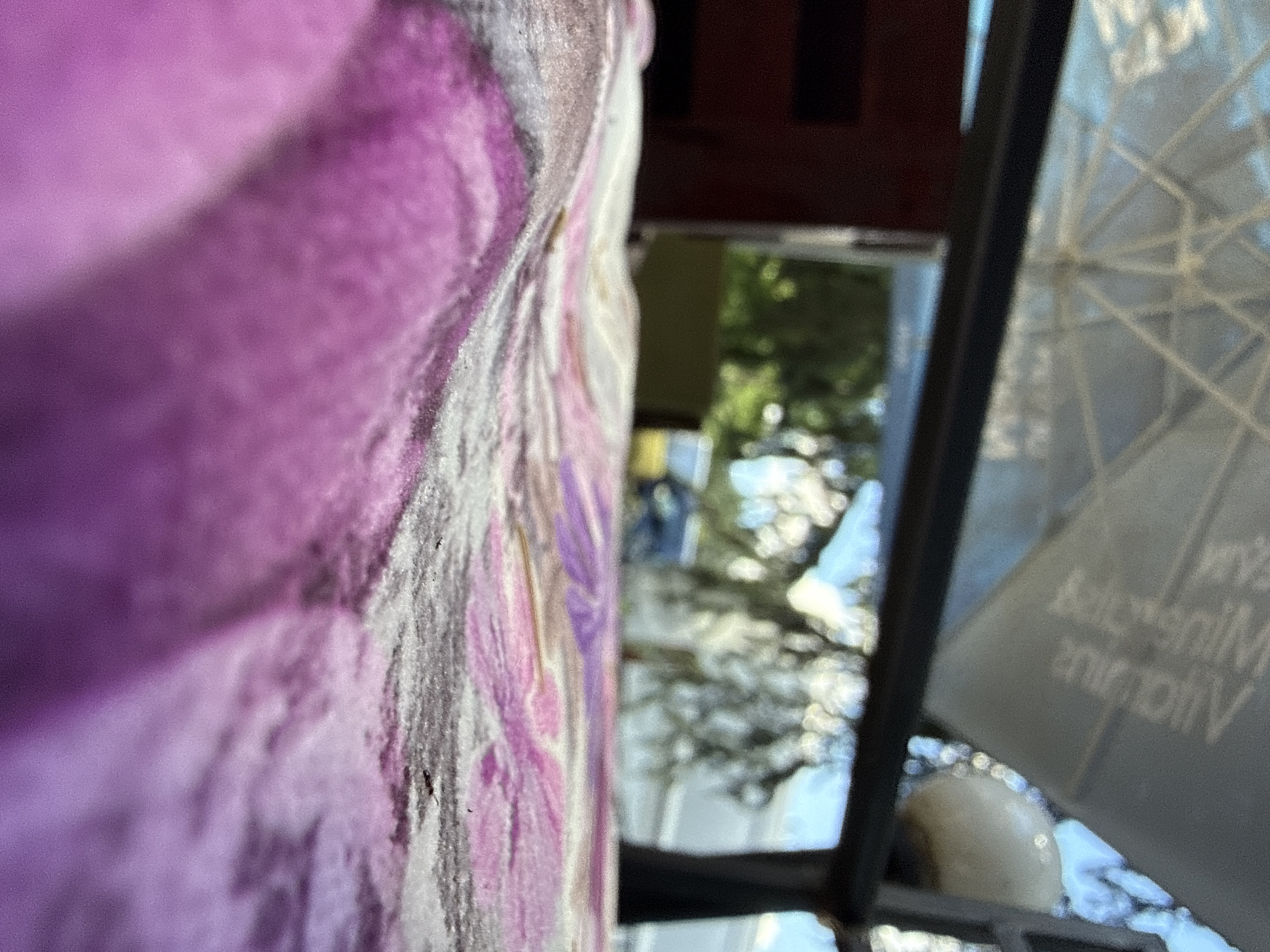 The image captures a close-up, low-angle view of a patterned fabric with shades of pink and purple. The fabric appears to be a tablecloth or blanket, with folds and creases creating a textured surface. In the background, a blurry scene of greenery, trees, and a partially visible structure with a geometric roof is discernible. The overall impression is one of an outdoor setting, possibly a garden or patio, with the foreground fabric dominating the composition. The location in Varna, Bulgaria, is not identifiable from the visual elements present.
