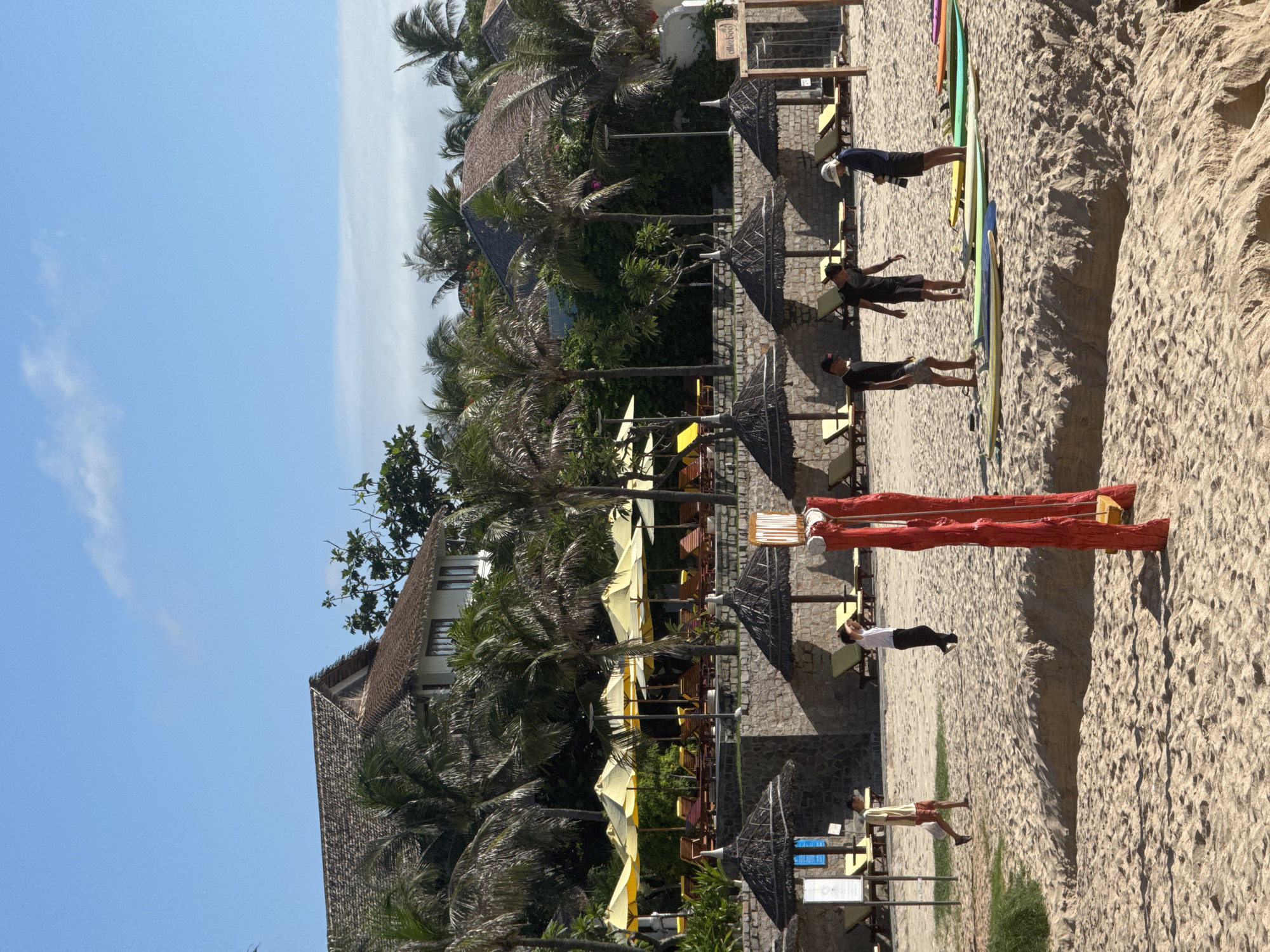 In Phan Thiết, Vietnam, this image captures a sandy beach scene on a sunny day. In the foreground, the sand is rippled, with a steep bank to the left. A red swing set with a wooden seat stands prominently. Further back, several people are gathered. Three young men, dressed in t-shirts and shorts, stand near a collection of surfboards lined up on the sand. One individual, wearing a white hat and dark shorts, appears to be taking a photograph or examining something on a surfboard. To the left, a young man in a light-colored shirt and shorts walks away from the camera.
In the middle ground, a stone wall separates the beach from a raised area with thatched umbrellas and rows of yellow lounge chairs. Beyond this, palm trees and lush green foliage obscure a resort with buildings that have thatched roofs. The sky is a clear, bright blue with a few wispy clouds. The overall atmosphere suggests a relaxed beach resort environment.