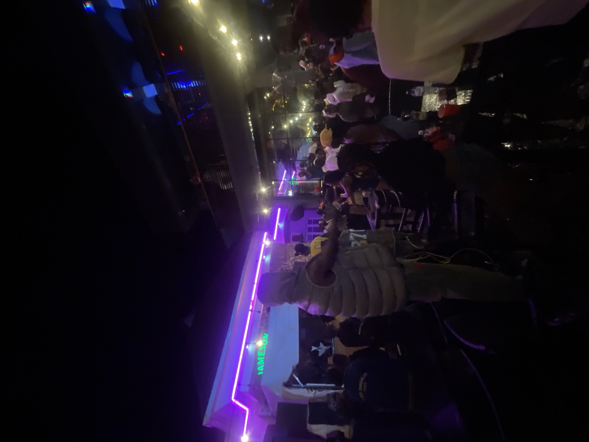 A nighttime scene showing a crowd of people gathered inside or in front of a building. The building has signage with the word "JAMESON" illuminated in green. The scene is lit with a combination of purple neon lights, string lights, and other building lights. The location is likely in Lokoja, Nigeria.