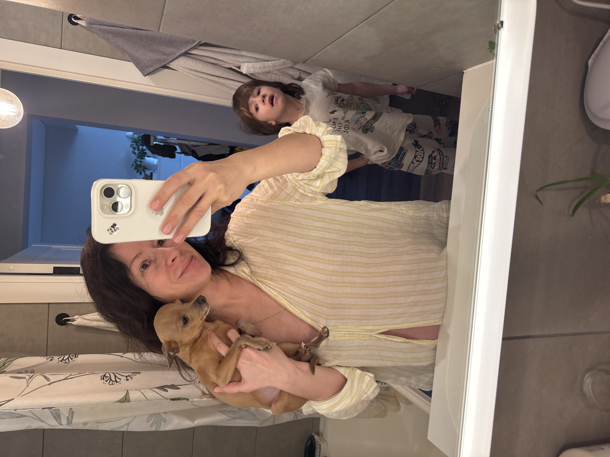 A woman is taking a selfie in a bathroom mirror, holding a small tan dog. A child is visible in the mirror behind her. The bathroom has light-colored tile walls, a gray towel hanging on a rod, and a shower curtain with a botanical pattern. The location is Chaikovsky, Russia.