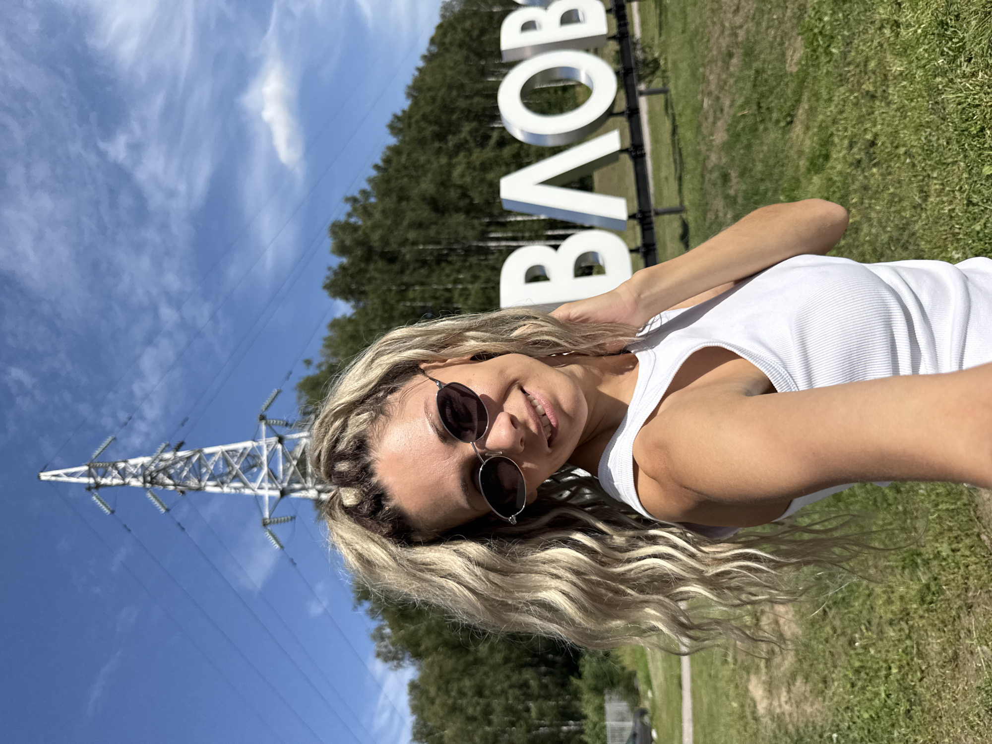 A woman with blonde hair styled in a high ponytail and sunglasses takes a selfie outdoors. She is wearing a white tank top. In the background, large white letters spell out "ВЛОВО". A tall electrical pylon stands behind her, with power lines stretching across the sky. A line of trees forms the horizon, and a grassy field with some dirt patches is visible in the foreground. The location is identified as Rusanovo, Russia.