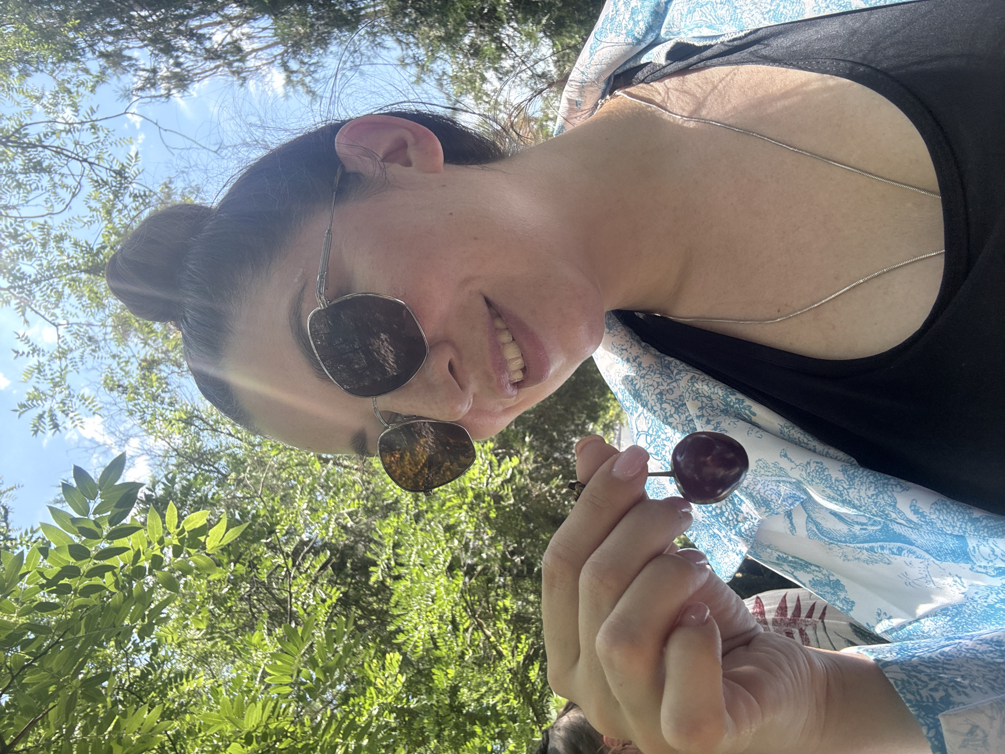 A low-angle selfie shows a woman in sunglasses holding two cherries. She has her hair in a bun and is wearing a black top with a blue and white patterned scarf draped over her shoulders. Her mouth is slightly open, revealing her teeth, and she is smiling. The background is filled with green foliage and a bright blue sky, suggesting an outdoor setting during the daytime. The lighting indicates it is sunny. The context places this image in Odesa, Ukraine.