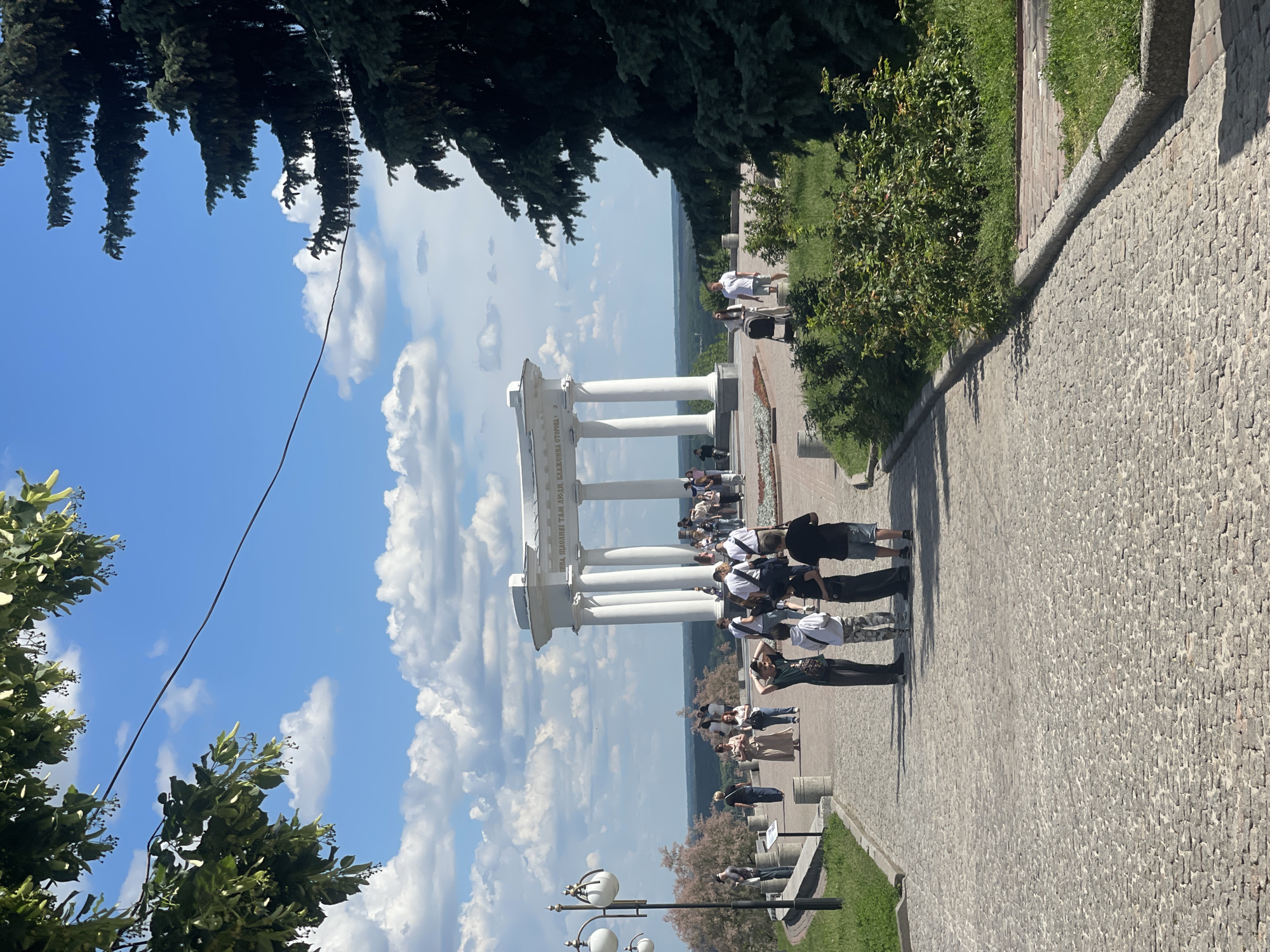 The image depicts a vibrant outdoor scene on a clear, sunny day, likely at a scenic overlook or promenade. A prominent white, classical-style rotunda, featuring numerous columns supporting an architrave, is positioned on a raised, paved platform. This rotunda serves as a focal point, with many people gathered around it, some observing the distant view, while others walk along the wide, cobblestone pathway.
The pathway extends from the foreground, curving past the rotunda, and is flanked by neatly maintained green bushes and grassy areas on the right. In the background, a vast, hazy green landscape stretches to the horizon, suggesting a wide valley or river plain. Above the scene, dark green coniferous trees create a shaded canopy, contrasting with the bright blue sky dotted with fluffy white cumulus clouds. A thin black wire is visible crossing the sky in the upper left portion of the frame.
The activity primarily involves people strolling, sightseeing, and congregating in small groups, indicative of a popular tourist spot or public park. Strong sunlight casts distinct shadows, suggesting the time of day is around midday or early afternoon. Visible text on the frieze of the rotunda is in Cyrillic, reading "УБЛАЖЕНА СТОЯЛИ СТОРОЙ," which translates approximately to "Blessed stood in a row." The overall mood is one of relaxed enjoyment amidst a picturesque setting.