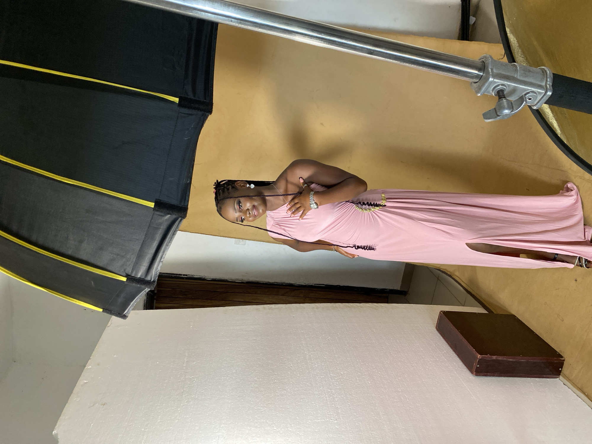 The image depicts a young woman posing for a photoshoot indoors. She is wearing a long, light pink, sleeveless dress with a high slit on the side. The dress has decorative embellishments around the waist. Her hair is styled in braids and she is wearing earrings and a watch. She is smiling and looking towards the camera, with her hands placed on her chest.
The setting appears to be a studio with a warm yellow backdrop. A large, black photographic umbrella with yellow accents is positioned to the left of the subject, suggesting professional lighting equipment is in use. A metal stand with a clamp is also visible, likely part of the lighting setup. In the foreground, a white surface, possibly a backdrop or a large styrofoam board, dominates the lower portion of the image. A dark brown, rectangular box or case rests on this white surface.
The image is captured in portrait orientation, with the subjects and scene rotated 90 degrees counter-clockwise. The lighting suggests an indoor environment, possibly during the day. The overall mood is positive, with the subject appearing confident and happy. No visible text is present in the image. The location context is Anguldi, Nigeria.