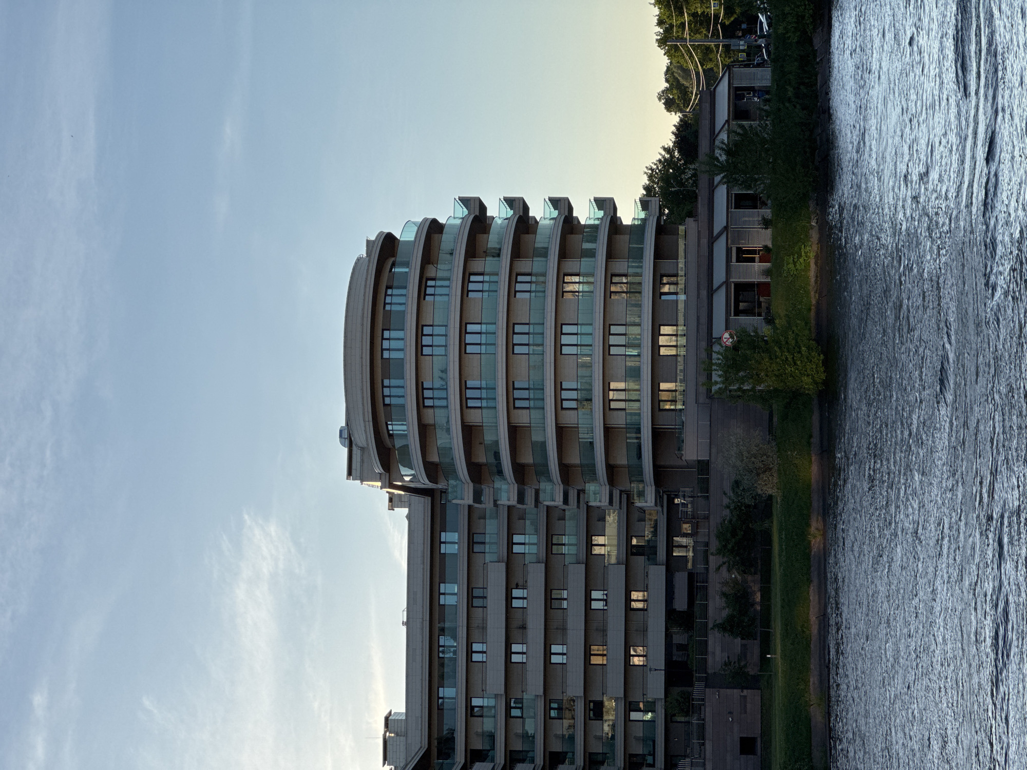 This is a medium shot, eye-level photograph of a modern, multi-story building with a curved facade, situated along a body of water. The building, constructed from a light-colored stone or concrete, features numerous windows and balconies with glass railings. The sunlight, casting a warm glow, illuminates the facade and reflects off the water. Trees and vegetation line the bank of the water in front of the building, suggesting a park-like setting. The sky above is a pale blue with wispy clouds, indicative of a clear day. In the background, more trees and some utility poles are visible. The scene is devoid of any people or vehicles. This building is the residential complex "Ohta Modern" located on the Ohta River in St. Petersburg, Russia.