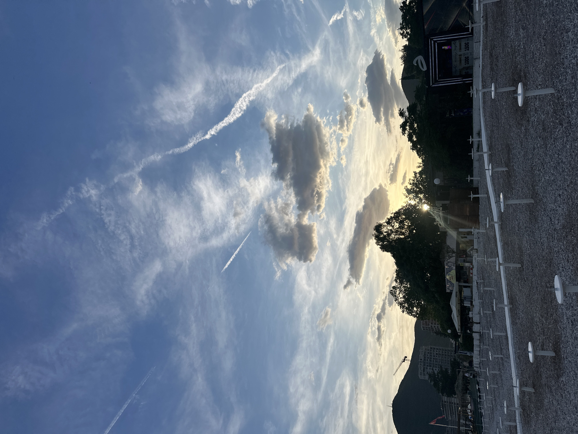 The image shows a view in Budva, Montenegro. The sky is predominantly blue, with scattered cirrus clouds and contrails from airplanes. The sun is partially obscured by clouds and trees, casting a warm glow. In the foreground, there is a gravel area with evenly spaced white posts. In the background, there are trees, buildings, and a mountain. A construction crane is also visible. The image appears to be taken in the late afternoon or early evening.