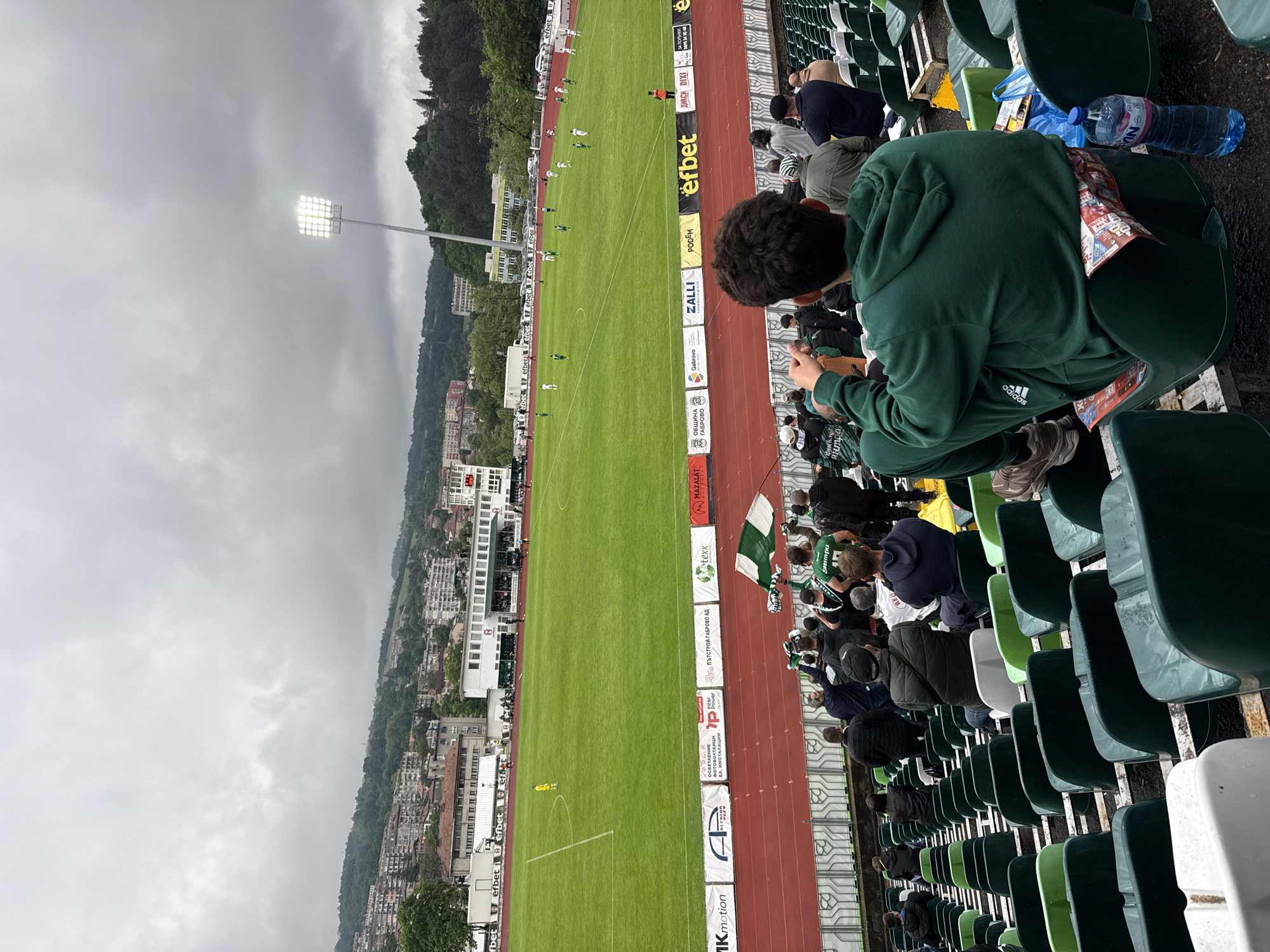 The image shows a soccer game being played in Gabrovo, Bulgaria. The perspective is from the stadium stands, with rows of green and white seats visible. A young person wearing a green Adidas tracksuit is sitting in the foreground, looking at a phone. The soccer field is in the middle, with players in green uniforms. A running track surrounds the field. Advertisements are displayed along the edge of the track. In the background, buildings of Gabrovo and a hill covered in trees are visible under a cloudy sky. A large lighting structure is visible to the left of the soccer field. A Bulgarian flag can be seen on the stands in the middleground of the image.