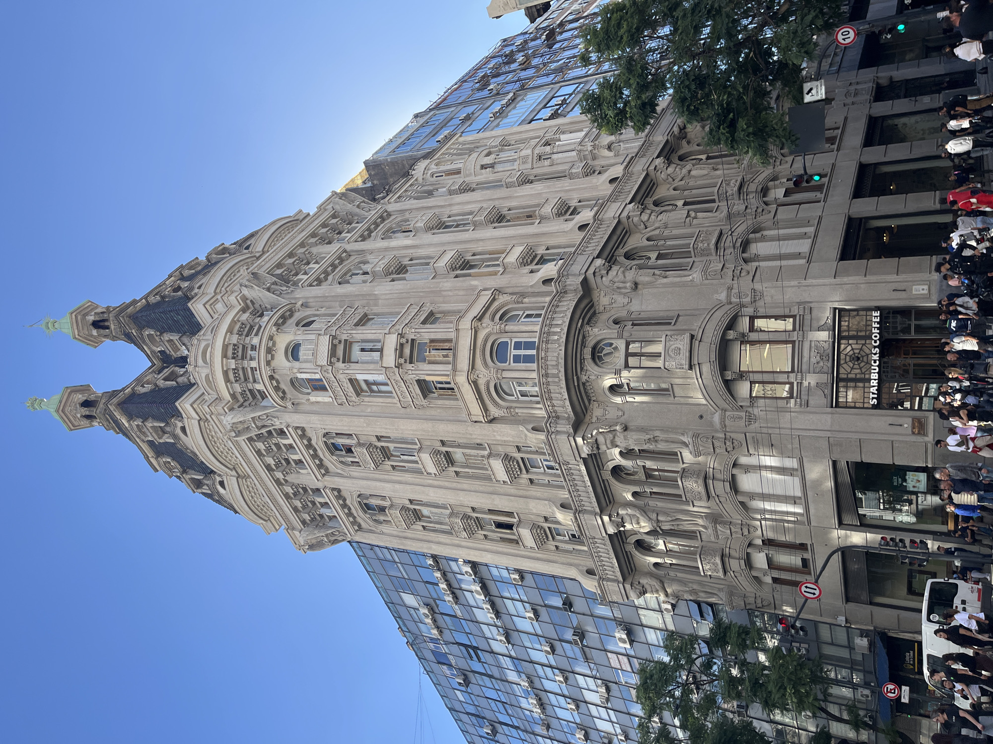 The image captures a bustling street scene in Buenos Aires, Argentina, under a clear, bright blue sky. A magnificent, ornate building with intricate architectural details dominates the left side of the frame. This historic structure features a light-colored, decorative stone facade with multiple stories, numerous arched windows, embellished balconies, and two distinct cupolas topped with green spires. The sunlight highlights its rich carvings and classical motifs.
Adjacent to this grand edifice, a modern glass-fronted building rises, reflecting the sky and surroundings. Numerous air conditioning units are visibly mounted on its exterior. A vibrant green tree partially frames the right side of the image, providing a natural element amidst the urban landscape.
At street level, the scene is lively with pedestrians. Many people are seen walking or standing, creating a sense of urban activity. A "STARBUCKS COFFEE" shop is prominently located on the ground floor of the ornate building, attracting some of the crowd. A white van is partially visible on the street, alongside various street signs. These include circular red signs displaying "10" and "11," likely indicating speed limits or route numbers, and a blue circular sign with a red diagonal line and an "E," signifying a no-parking zone. Inside a shop window, the word "DULCE" is visible, and "PRE" can be seen on a person's shirt.
The strong sunlight and clear sky suggest it is daytime, likely morning or early afternoon. The photograph portrays a dynamic metropolitan environment, characterized by a striking juxtaposition of historic European-influenced architecture and modern city elements.