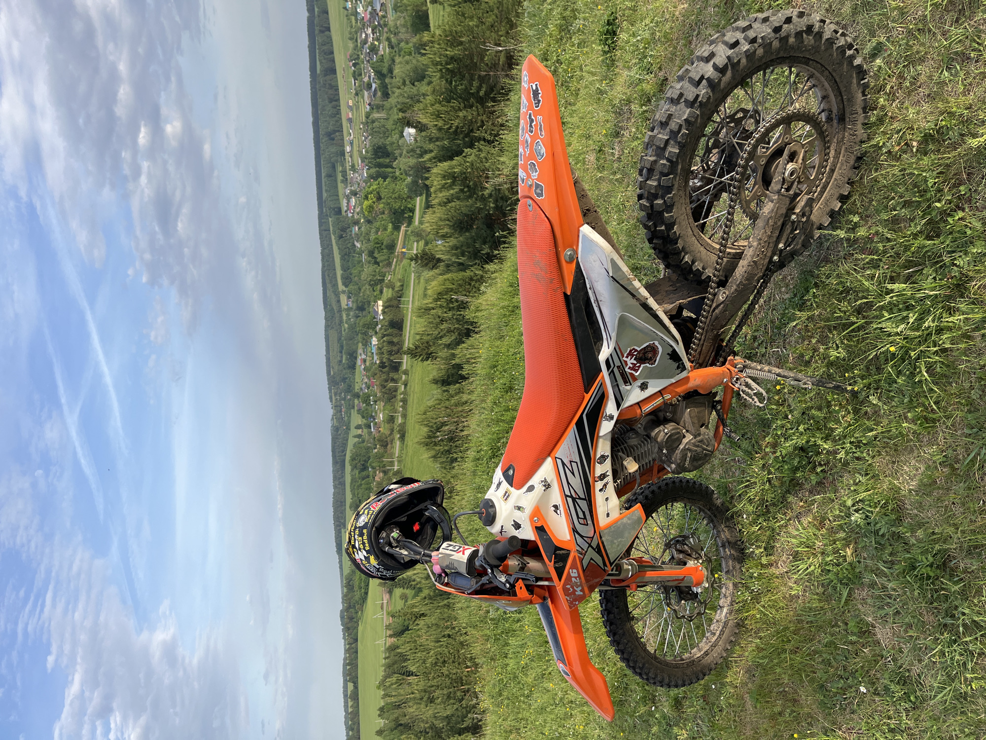 The image depicts a dirt bike resting on its side in a grassy field. The bike is predominantly orange and white with some black accents. The seat is orange and the bike has stickers on the rear fender. A black helmet with colorful decals rests on the handlebars. The background features a landscape of green fields, trees, and a village or town in the distance under a partly cloudy sky. The location appears to be rural, with an expansive view.