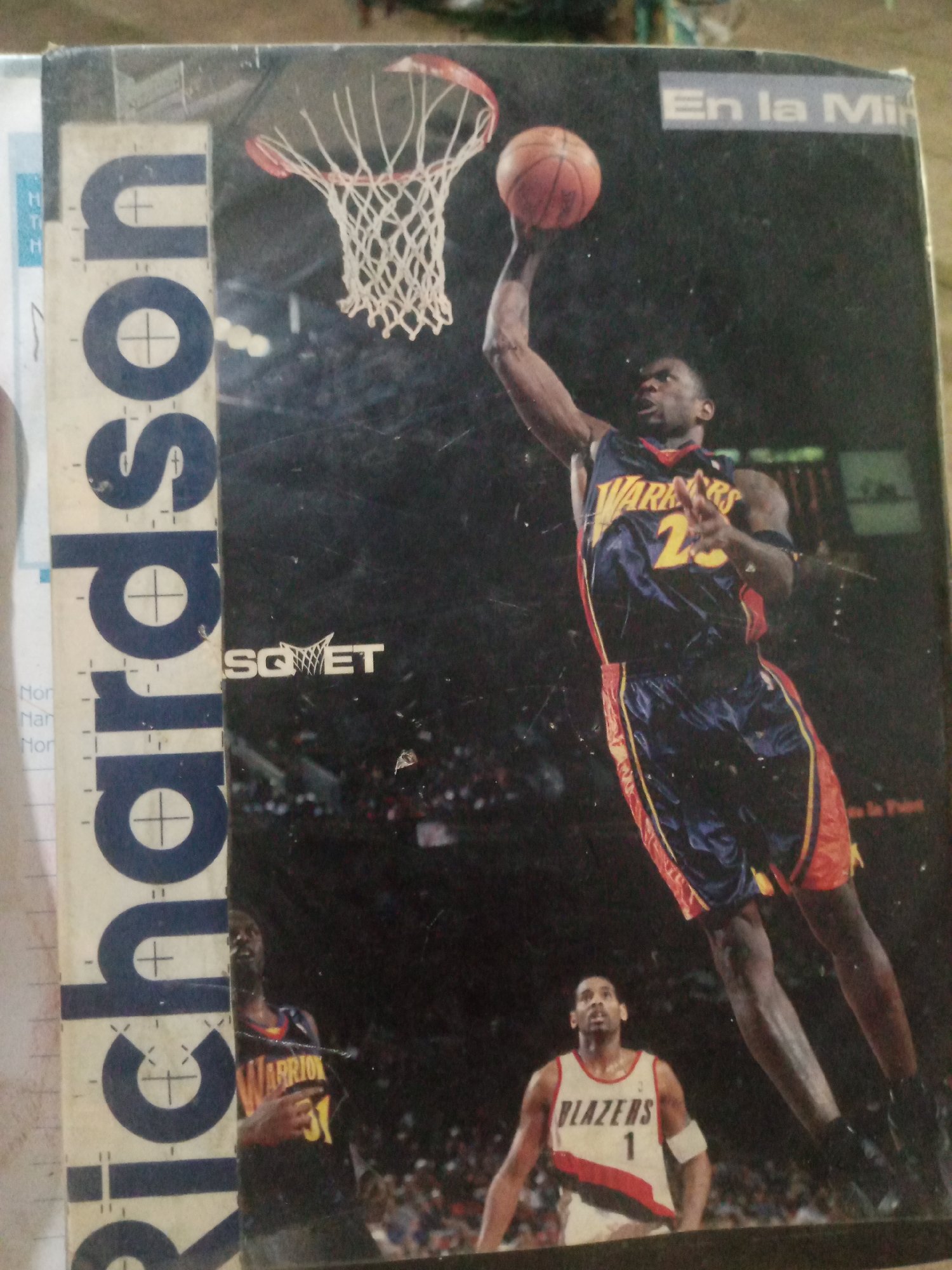 The image appears to be a photograph of a magazine cover or a poster. The main subject is a basketball player in mid-air, attempting a dunk. He is wearing a blue and gold uniform with the number 23. Another basketball player, wearing a white and red uniform with the number 1 and the word "BLAZERS," is looking up at the dunking player. The background is dark and appears to be a basketball arena with spectators. There is text at the top right that reads "En la Mi". On the left side of the image, there is vertical text that appears to be a name or title, partially obscured. There are also some markings and a drawing of a basketball hoop. The image quality is somewhat degraded with scratches and dust. There is no identifiable information within the image that would suggest a location in Santa Ana, Venezuela.