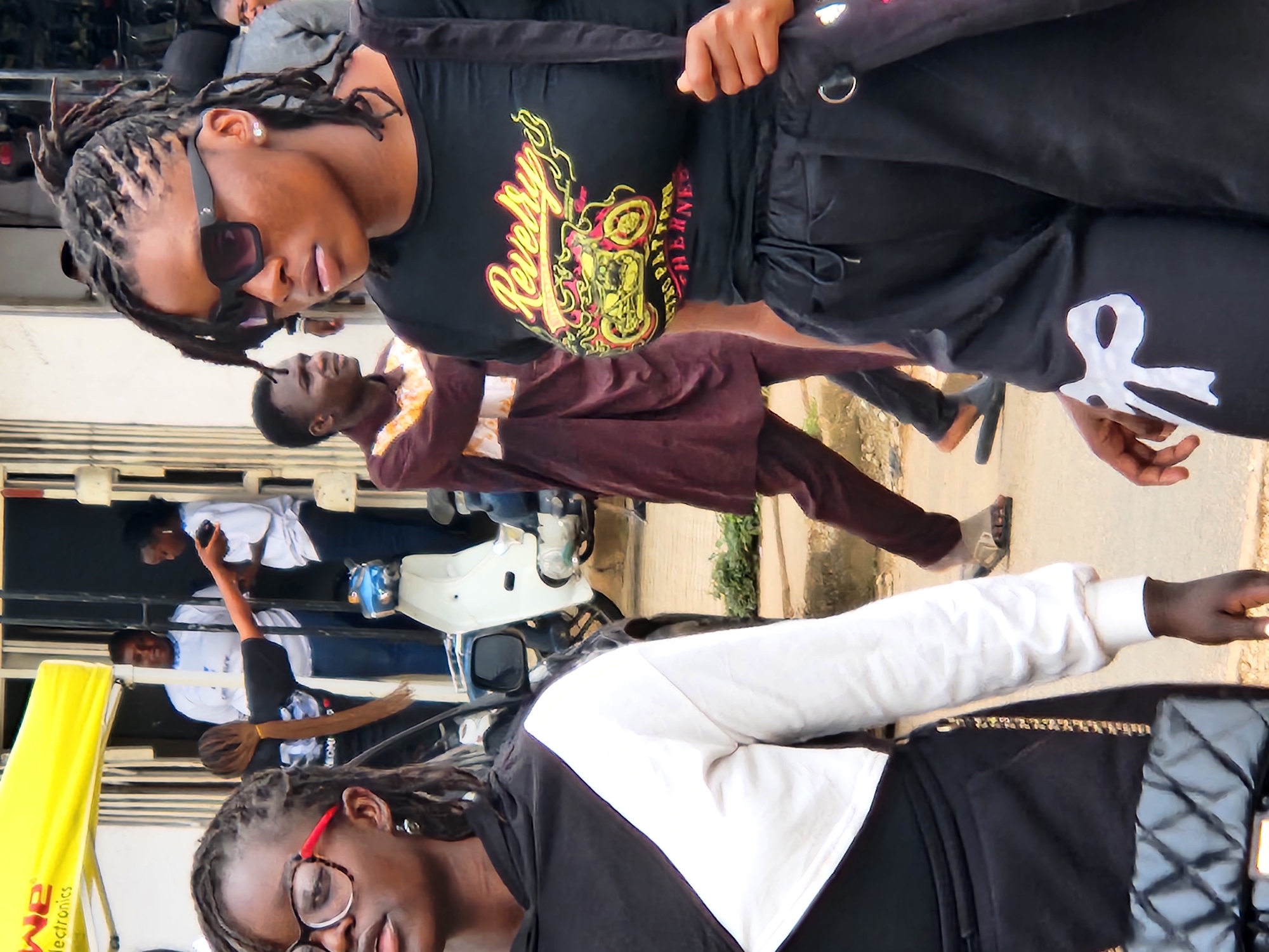 The image, taken in an outdoor urban environment, captures a group of people in motion. In the foreground, a young woman with dark skin and braided hair, wearing sunglasses, a black t-shirt with a graphic print, and black pants, is walking. She has a black purse slung over her shoulder. To her left, another young woman with dark skin, wearing glasses and a black and white hooded top, is also walking. In the background, a man in a brown tunic and pants walks past. Other individuals are visible in the background, including two women in white tops and dark pants, and a person in a grey jacket. A white scooter and a partial view of a yellow awning are also present. The scene appears to be a busy street or market area.