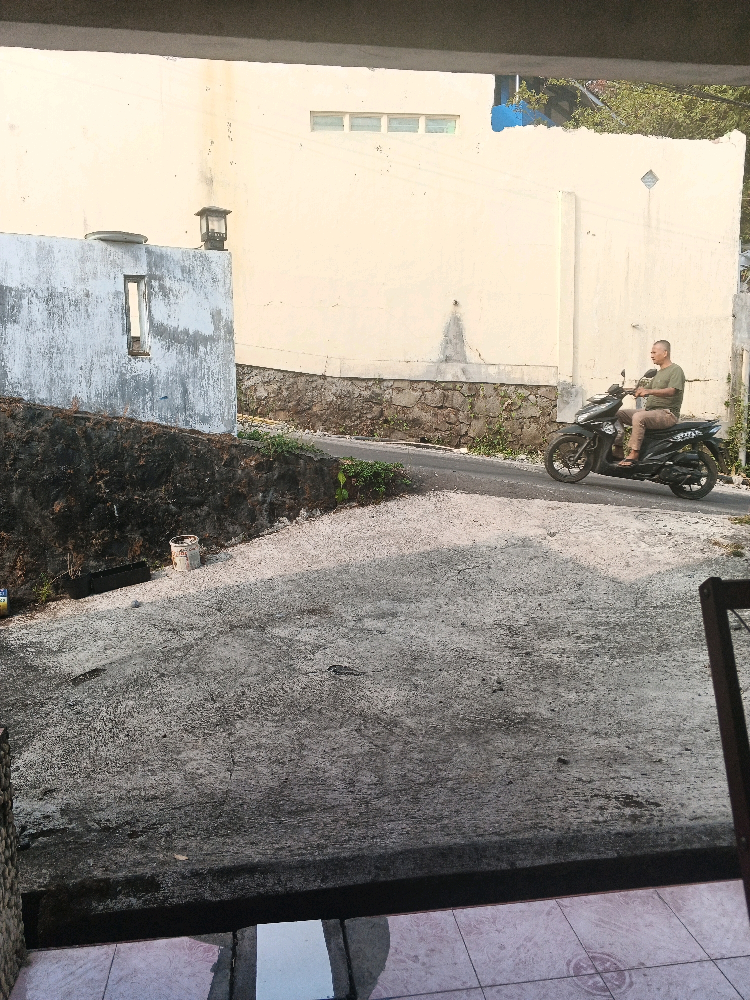 A bald man wearing a green short-sleeved shirt and brown pants rides a black motorcycle on a paved road that slopes upward. The man is looking to his right. The road curves slightly to the right. To the left of the road is a dark, rough-textured wall covered in moss and dirt, with a white building above it that has a small, rectangular opening. To the right of the road is a light yellow wall with a few small, rectangular windows. There is a stone wall separating the yellow wall from the road at the bottom. The scene appears to be in Keji, Indonesia, based on the typical architecture and environment. The lighting suggests it is late afternoon or early morning, with shadows cast on the ground.
