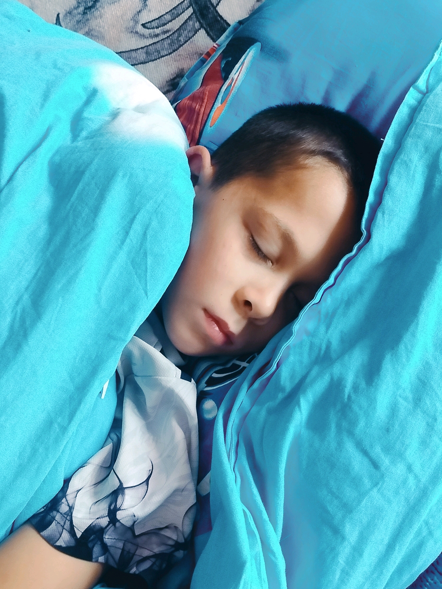 The image shows a close-up, overhead view of a young boy sleeping peacefully under a bright blue comforter. The boy's face is turned slightly to the right, with his eyes closed and his mouth slightly open. His dark hair is short and neatly combed. The comforter is pulled up around his head and shoulders, creating a sense of warmth and security. The background of the image is blurred, but it appears to be a bed with patterned pillows and blankets. There are no specific landmarks or details in the image that would indicate a location within the city of Көлкент, Kazakhstan. The scene is domestic and personal, focusing on the intimate moment of a child at rest.