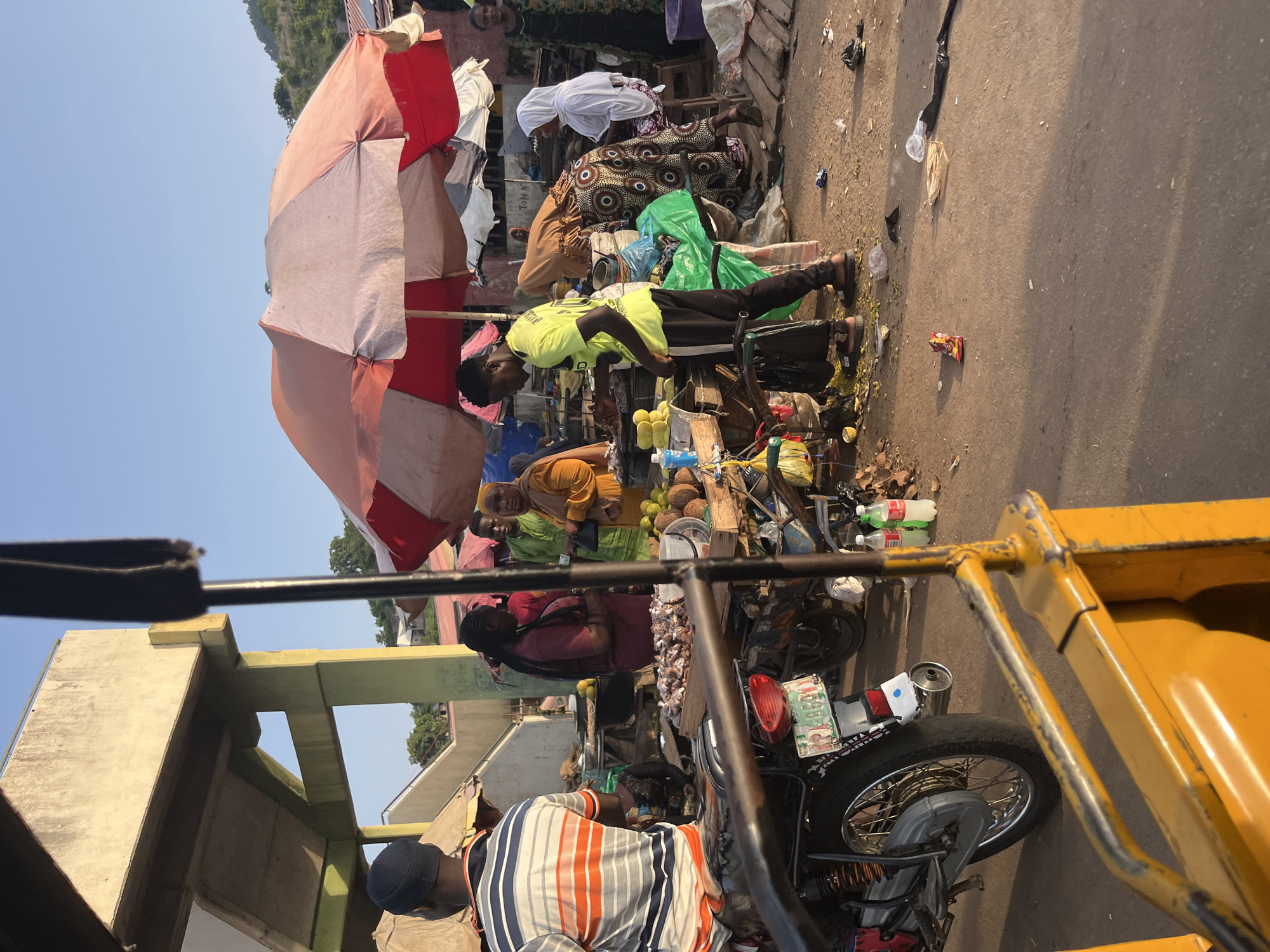 The image depicts an outdoor market scene in Unknown, Unknown. A man in a bright yellow shirt and black pants stands near a market stall, appearing to prepare goods for sale. He is surrounded by various items including coconuts, yellow fruits, and what looks like small dried fish or meat. A woman in a brown hijab and patterned dress is seated behind the stall. Another woman, wearing a white hijab and patterned skirt, sits on a stool nearby. In the foreground, a person wearing a striped t-shirt and a blue cap is seated on a motorcycle. The market appears to be bustling, with other individuals visible in the background, some tending to stalls and others browsing. Large umbrellas, red and pink, provide shade. The ground is paved and littered with some debris. The overall environment suggests a lively, daily market day.