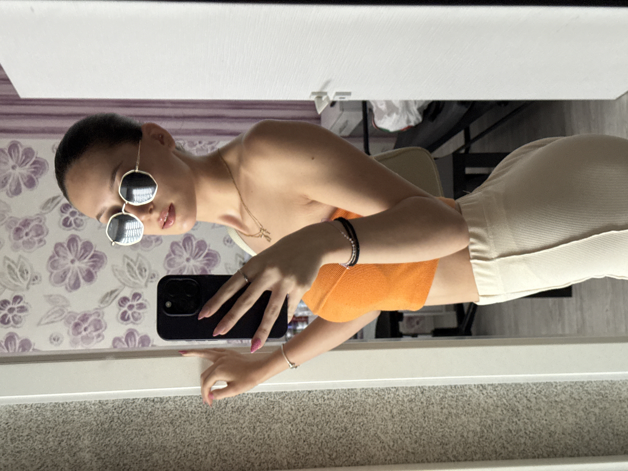 An unseen person is taking a selfie in a room, with a mirror reflecting their image. They are wearing a strapless orange top, beige ribbed pants, and round, gold-rimmed sunglasses. A light-colored purse is slung over their right shoulder. The background shows a wall with purple floral wallpaper and a white door. There is no indication of the city of Sofia, Bulgaria in the image.