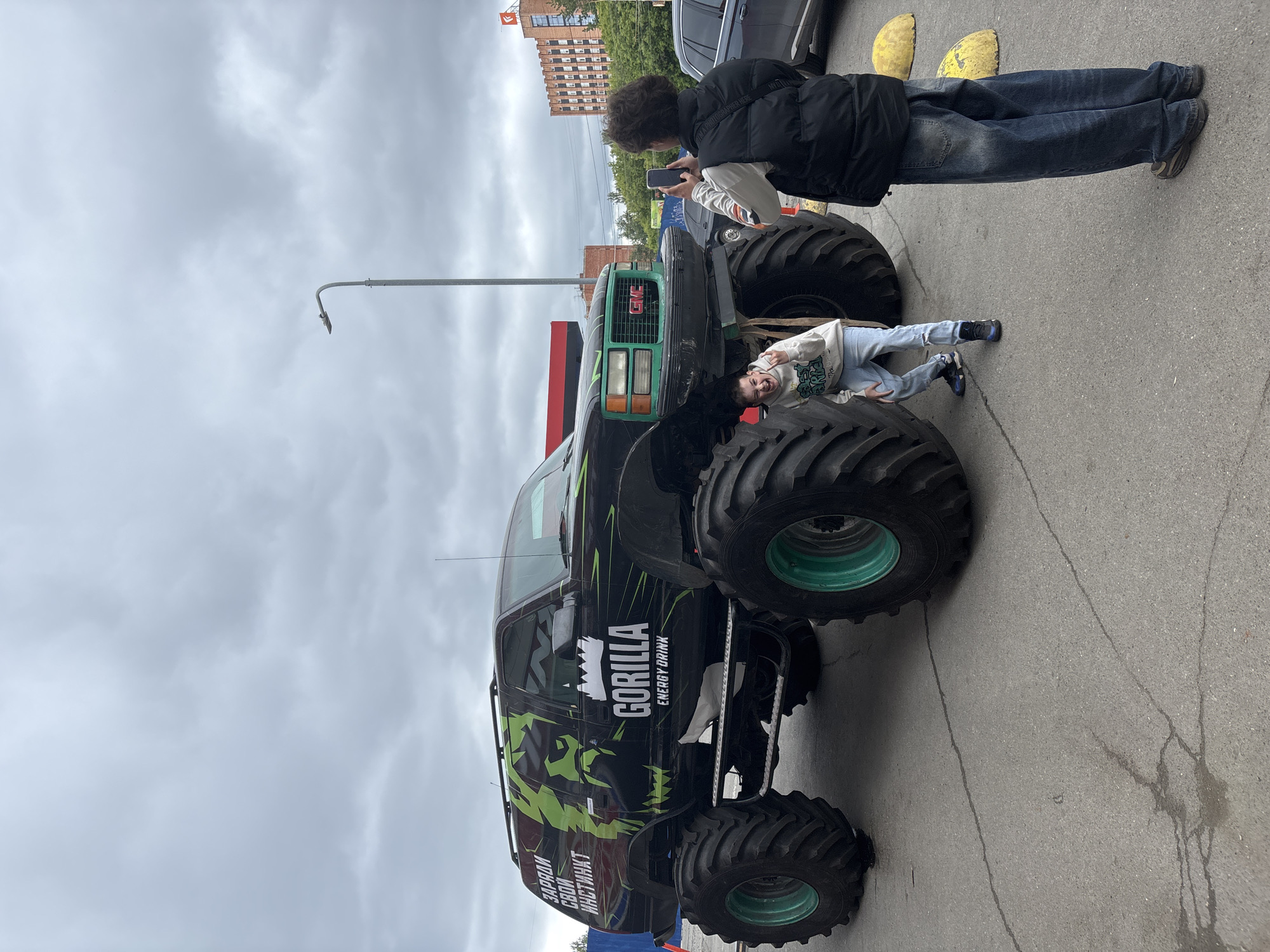 The image depicts an outdoor urban scene in Izhevsk, Russia, on an overcast day. Central to the frame is a black monster truck, modified from an older GMC SUV, featuring oversized black tires with prominent bright green rims. The truck's body is adorned with green and black graphics, including a stylized gorilla head, and the branding "GORILLA ENERGY DRINK." The Russian text "ЗАРЯДИ СВОЙ ИНСТИНКТ" (Charge Your Instinct) is visible on the rear side.
Two males are interacting with the vehicle. On the left, a young male, wearing a light-colored hooded sweatshirt with graphics, ripped light blue jeans, and dark sneakers with blue accents, crouches low between the front and rear tires on the driver's side. He faces the viewer with an open-mouthed expression of excitement or laughter, holding his left hand up in a gestural pose. On the right, a second male stands with his back mostly to the viewer, holding a black smartphone horizontally to photograph the first male and the truck. He wears a black puffy vest over a light-colored long-sleeved shirt with striped sleeves, loose-fitting faded blue jeans, and dark sneakers.
The ground is asphalt pavement with visible cracks and some damp spots. In the background, an urban environment is present, including a red structure possibly belonging to a gas station or commercial establishment on the left, and a dark-colored SUV parked on the right. Further back, trees partially obscure a multi-story, reddish-brown brick building with numerous windows, topped by a red flag featuring a white logo. A grey lamppost with a curved light fixture is visible on the right side, extending towards the overcast, heavily clouded sky. Two yellow, hemispherical concrete bollards are situated on the ground to the right of the monster truck.
