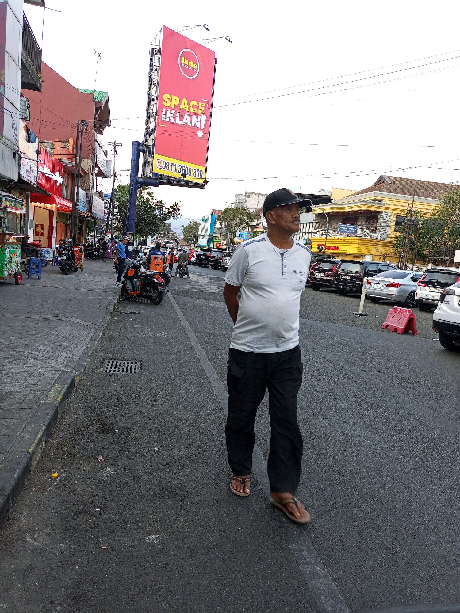 The image depicts a street scene in Batu, Indonesia. In the foreground, a man wearing a light-colored, short-sleeved shirt, black pants, and sandals stands with his hands behind his back, looking to his left. He is wearing a black baseball cap. To his right, the street is paved with asphalt and a white line marks a lane. Several cars are parked on the right side of the street. Red and white traffic barriers are visible near the parked cars.
To the left of the man, a sidewalk is present, bordered by a curb. On the sidewalk, there are various street vendors and shops. A food cart with a green and white sign is visible. Several motorcycles are parked along the sidewalk. Buildings line the left side of the street, featuring storefronts and upper-story windows.
Dominating the background is a large red billboard with "Jade Indopratama" at the top, followed by "SPACE IKLANI" in large yellow letters, and a phone number at the bottom. The sky is overcast. The overall scene suggests a bustling commercial area.