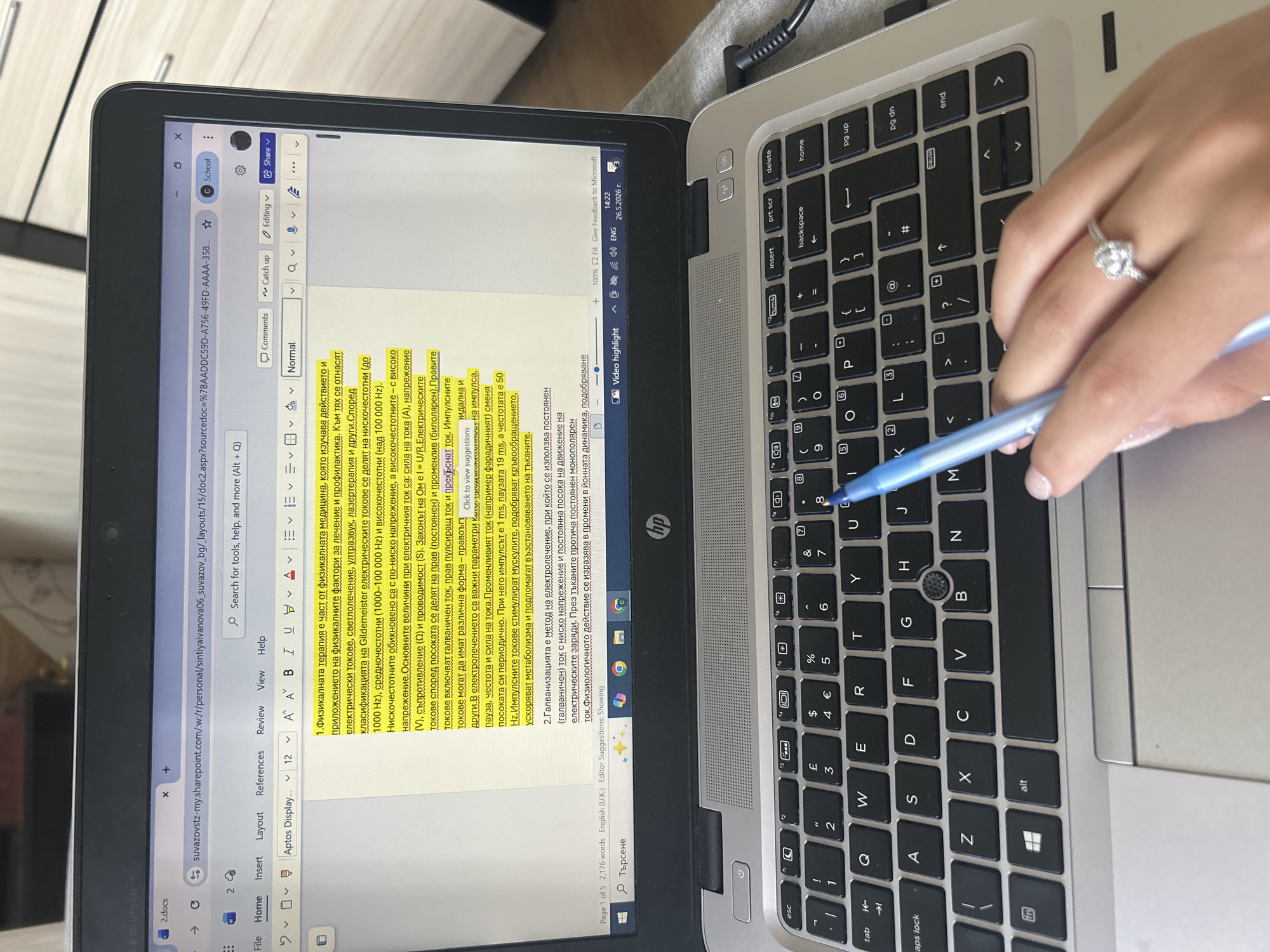 Here's a concise description of the image:
A person is shown using a laptop. The laptop is open, and the screen displays a document in a word processing program (likely Microsoft Word). The document appears to be in Bulgarian, containing text about physical therapy and electrical currents. The person is holding a blue pen near the keyboard. The keyboard is a standard QWERTY layout. The person is wearing a ring with a large stone. The laptop is resting on a gray surface. In the background, a piece of furniture with light wood is visible.