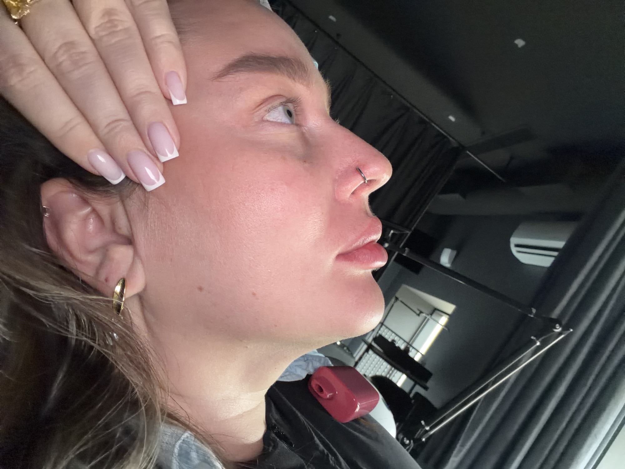 A light-skinned person, possibly female, is the central subject, viewed in profile from the left. They have blonde or light brown hair and fair skin. They have a silver nose ring and gold earrings. Their nails are manicured with a white French tip style.
The background appears to be an indoor space, possibly a salon or studio, given the presence of what appears to be a hair styling or camera lighting setup. A window and what appears to be an air conditioning unit are visible in the background. The location cannot be determined from the image.