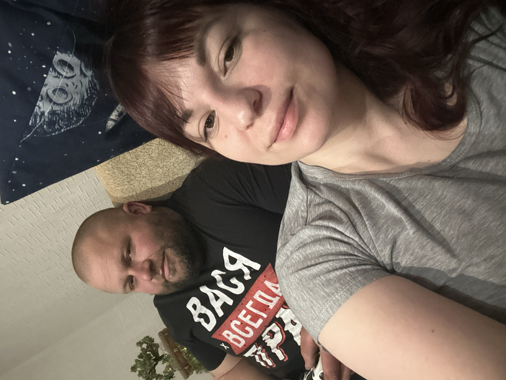A selfie shows a woman and a man lying on a surface covered with a blanket with a moon and star design. The woman has brown hair and is wearing a gray T-shirt. The man has a shaved head, a beard, and is wearing a black T-shirt with Cyrillic text. The location is unknown but may be in Фоки, Russia.