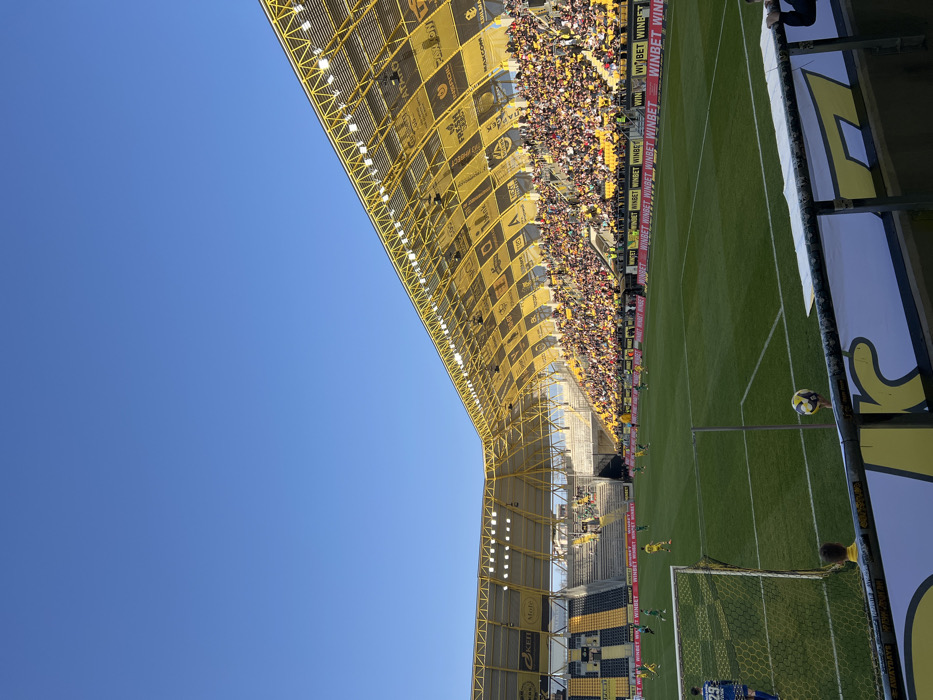 The image displays an outdoor football stadium, identified as Lokomotiv Stadium (Lauta Stadium) in Plovdiv, Bulgaria, under a clear blue sky during daylight hours. The perspective is low, positioned behind a goal on the left side of the frame, looking across the pitch towards the opposite main stand.
The stadium features a distinctive yellow metal roof structure, equipped with numerous floodlights, extending over the stands. This roof is patterned with black square advertising panels displaying various sponsor logos, including "WINBET," "FRESH," and "MAX.COM," some of which feature Cyrillic script. The main stand on the right is densely populated with spectators, many of whom are wearing yellow and black attire, consistent with the home team's colors. The visible seating in the stands alternates between black and yellow stripes.
On the well-maintained green football pitch, a match is in progress. Several players in bright green kits are visible on the left side of the field, while opposing players in yellow kits are visible on the right. A goalkeeper in a blue kit, wearing the number 29, stands near the foreground goal, which is white-framed with a net. One football is visible on the pitch near midfield. In the immediate foreground, a dark metal barrier with a white and yellow advertising banner for "WINBET" partially obstructs the view. Above this barrier, a person's hand is seen holding a second football. A person in a yellow shirt is partially visible below the barrier.